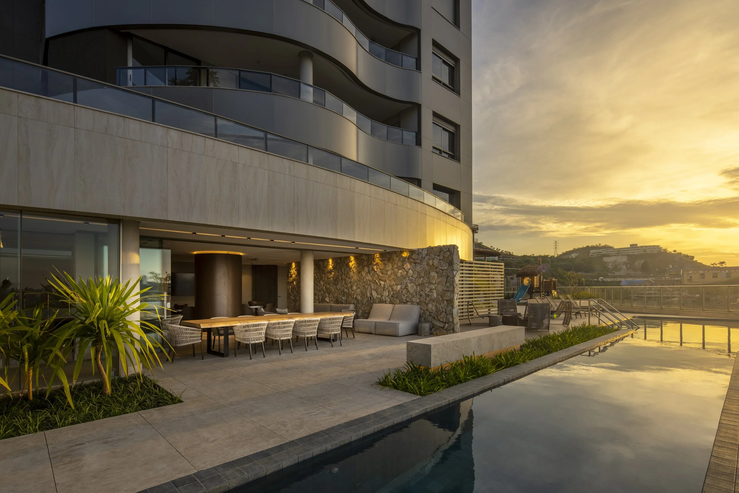 Vista de um prédio moderno ao pôr do sol, com varanda arredondada, fachada de Mármore e janelas grandes. Na área externa, há móveis de jantar, poltronas e uma piscina refletindo o céu.