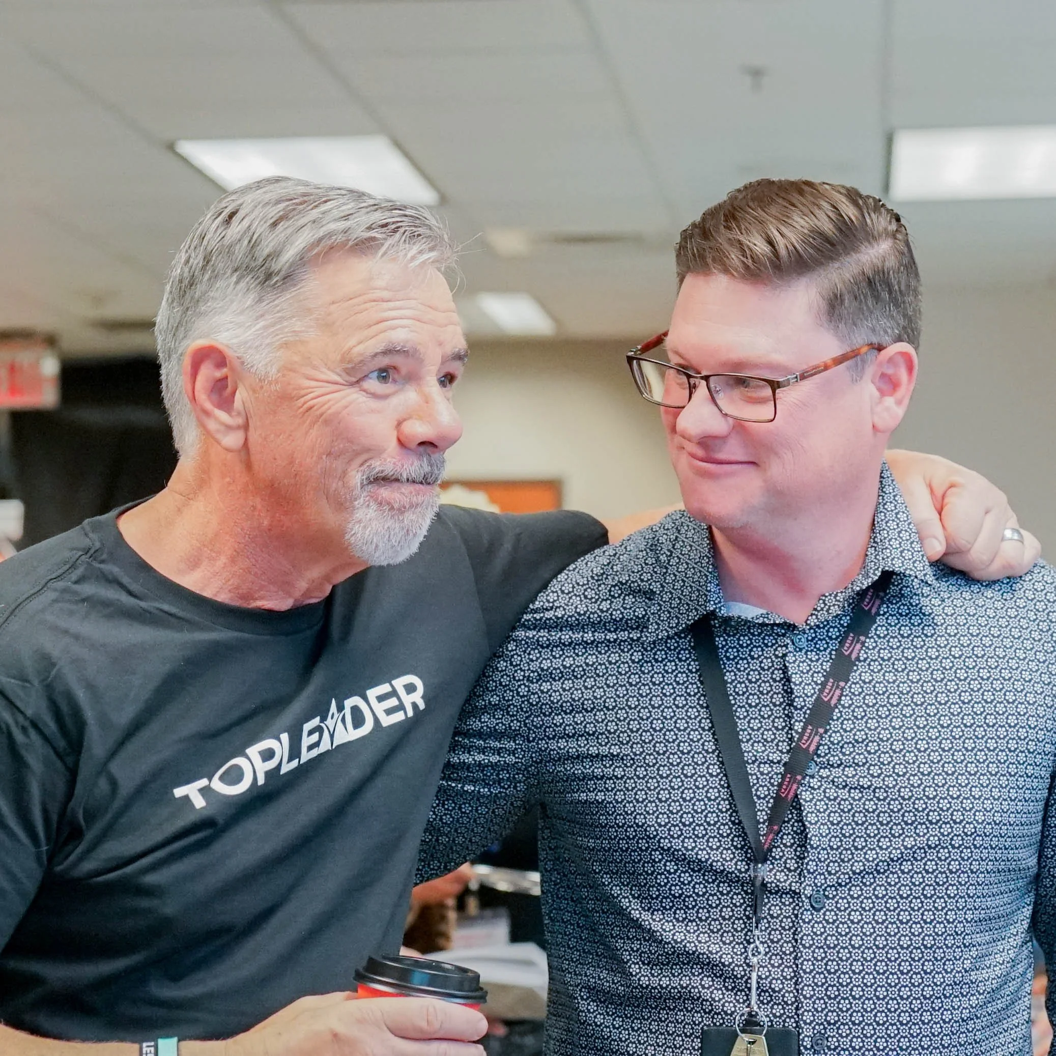 Two men having a friendly conversation, one with gray hair and beard wearing a black T-shirt with 'TOP LEADER' logo, the other with short brown hair and glasses wearing a patterned shirt, in an indoor setting.