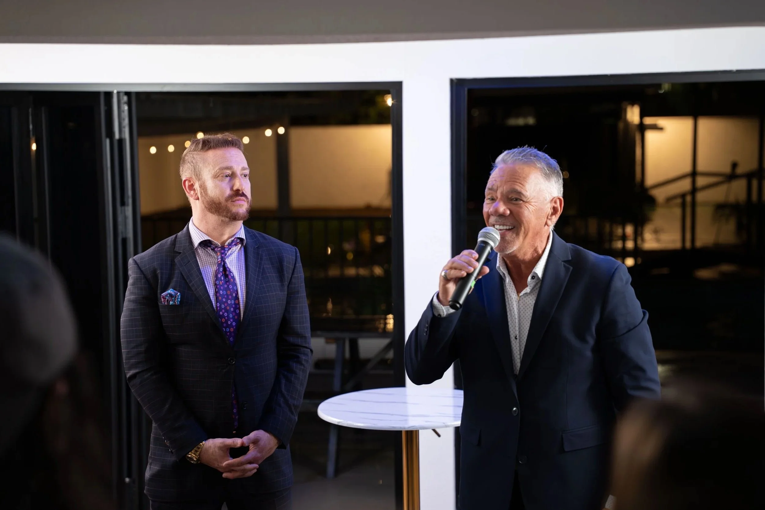 Two men in suits speaking at an indoor event, one holding a microphone and smiling, the other standing with hands clasped, with windows and outdoor lights in the background.