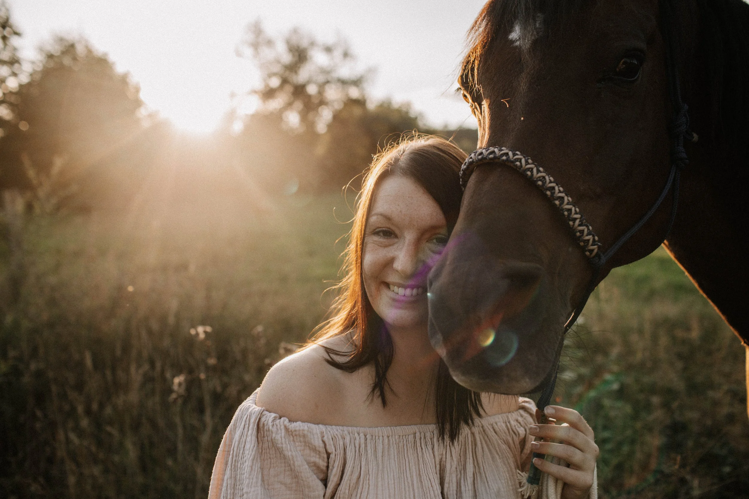 Eine Frau im beigen Kleid lächelt in die Kamera und steht neben einem braunen Pferd im Freien bei Sonnenuntergang.