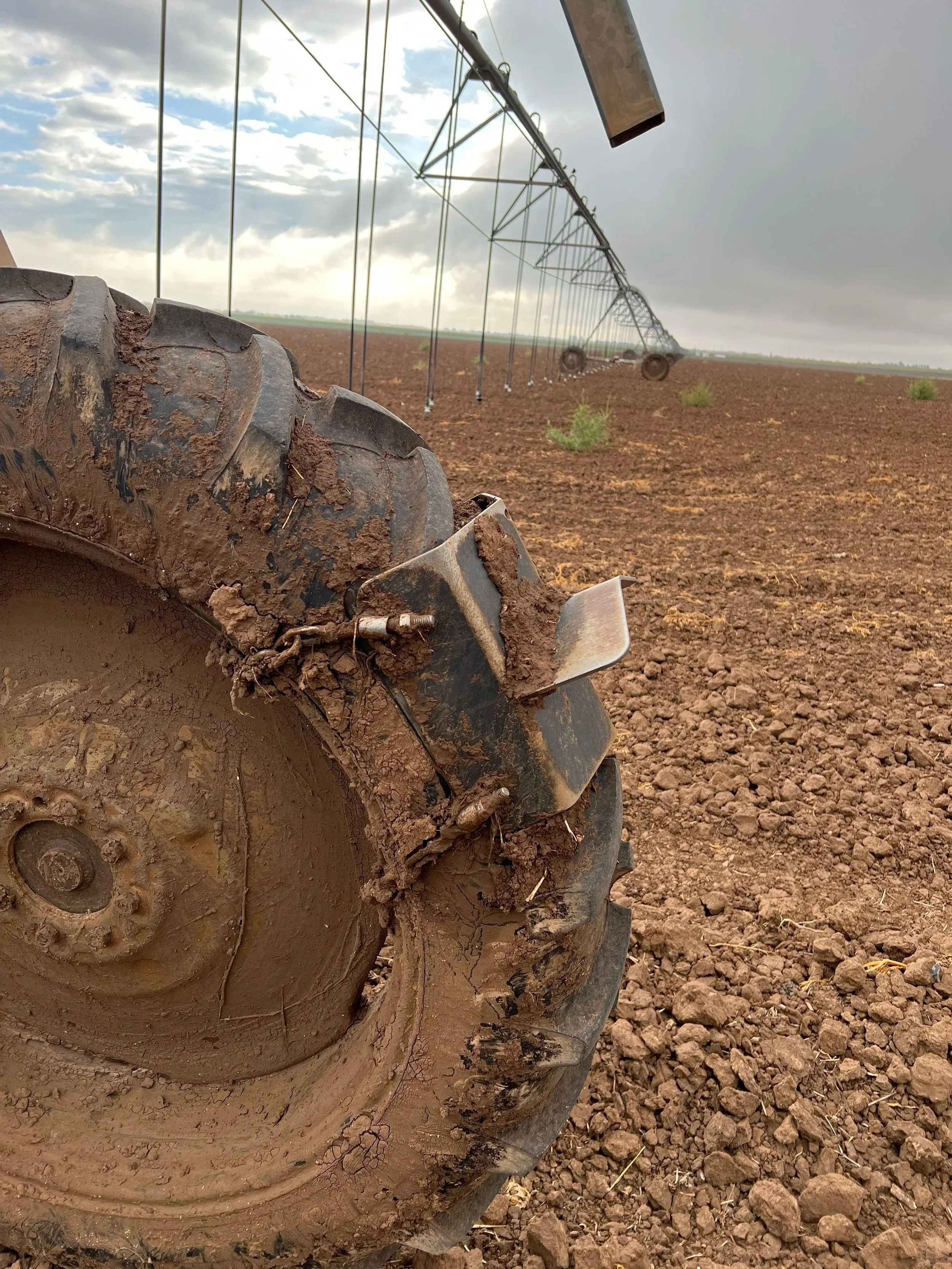 Close-up of a tractor tire with mud, in a plowed field during overcast weather, showing a pivot irrigation system stretching into the distance.
