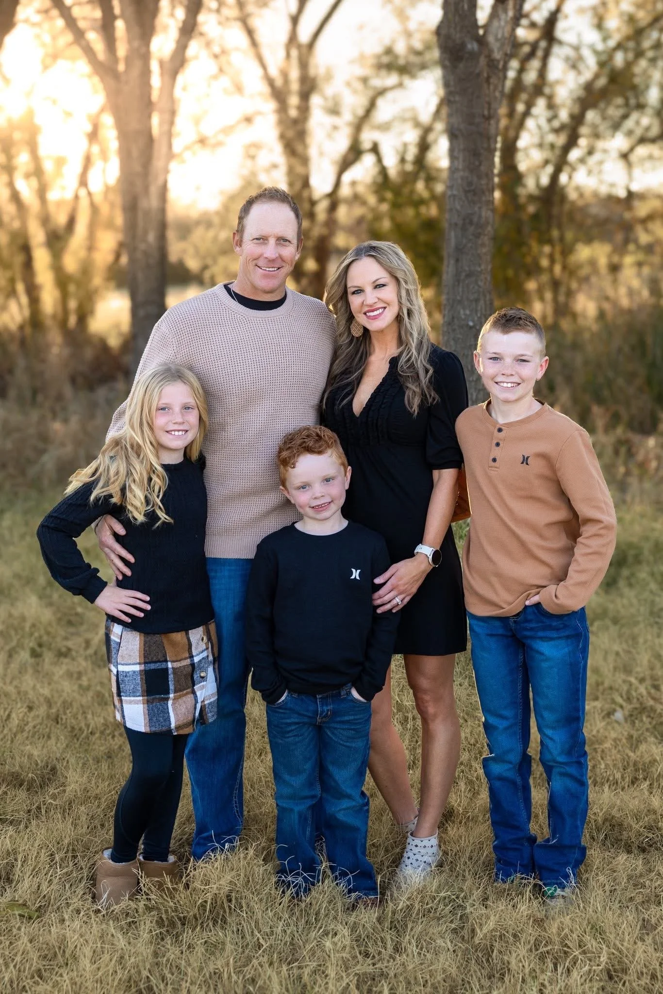 Family of five outdoors during fall sunset, standing on grass with trees in background, smiling for the camera.