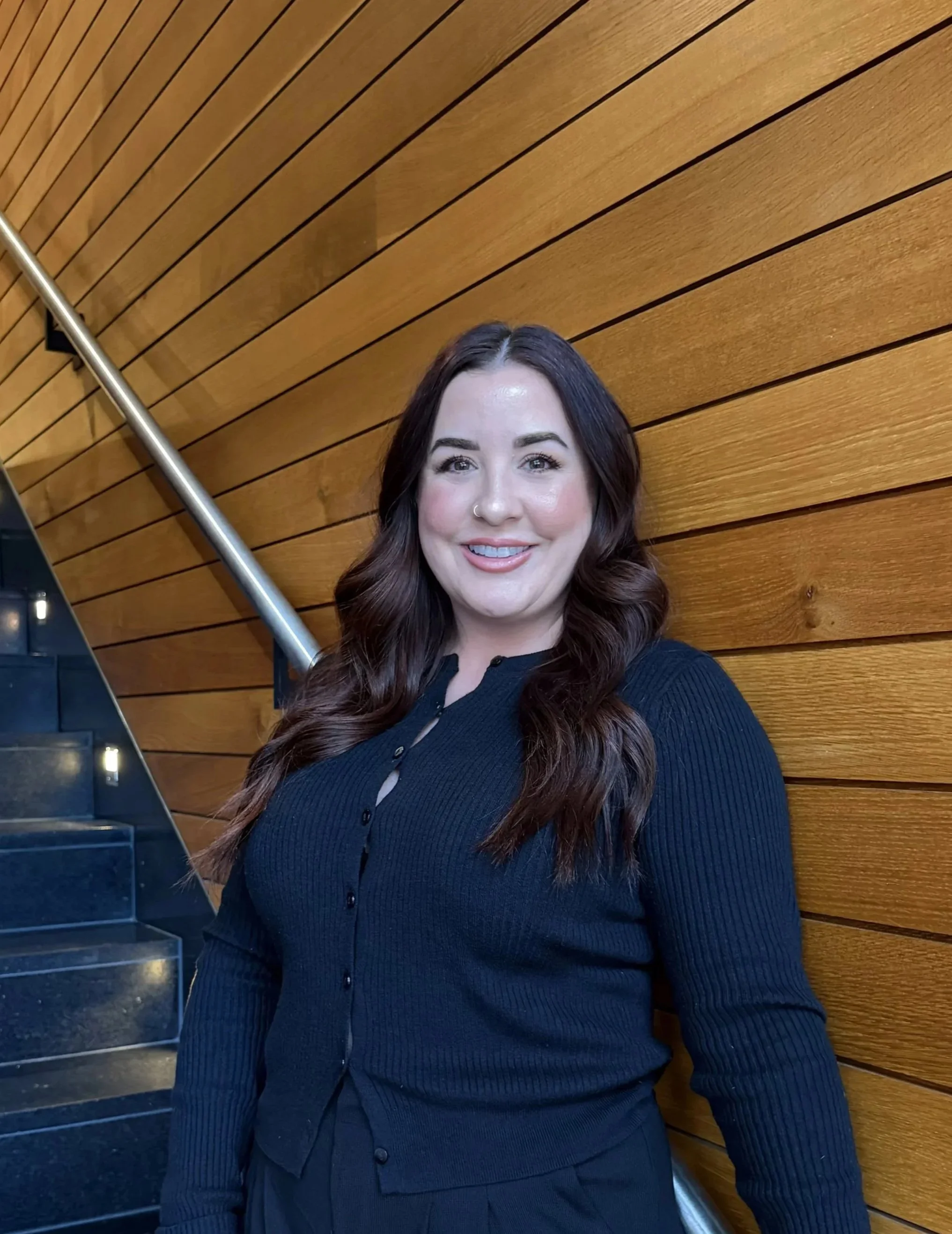 A woman with long wavy dark brown hair, wearing a black button-up sweater, smiling, positioned against a wooden paneled wall with a metal railing and stairs visible on the left.
