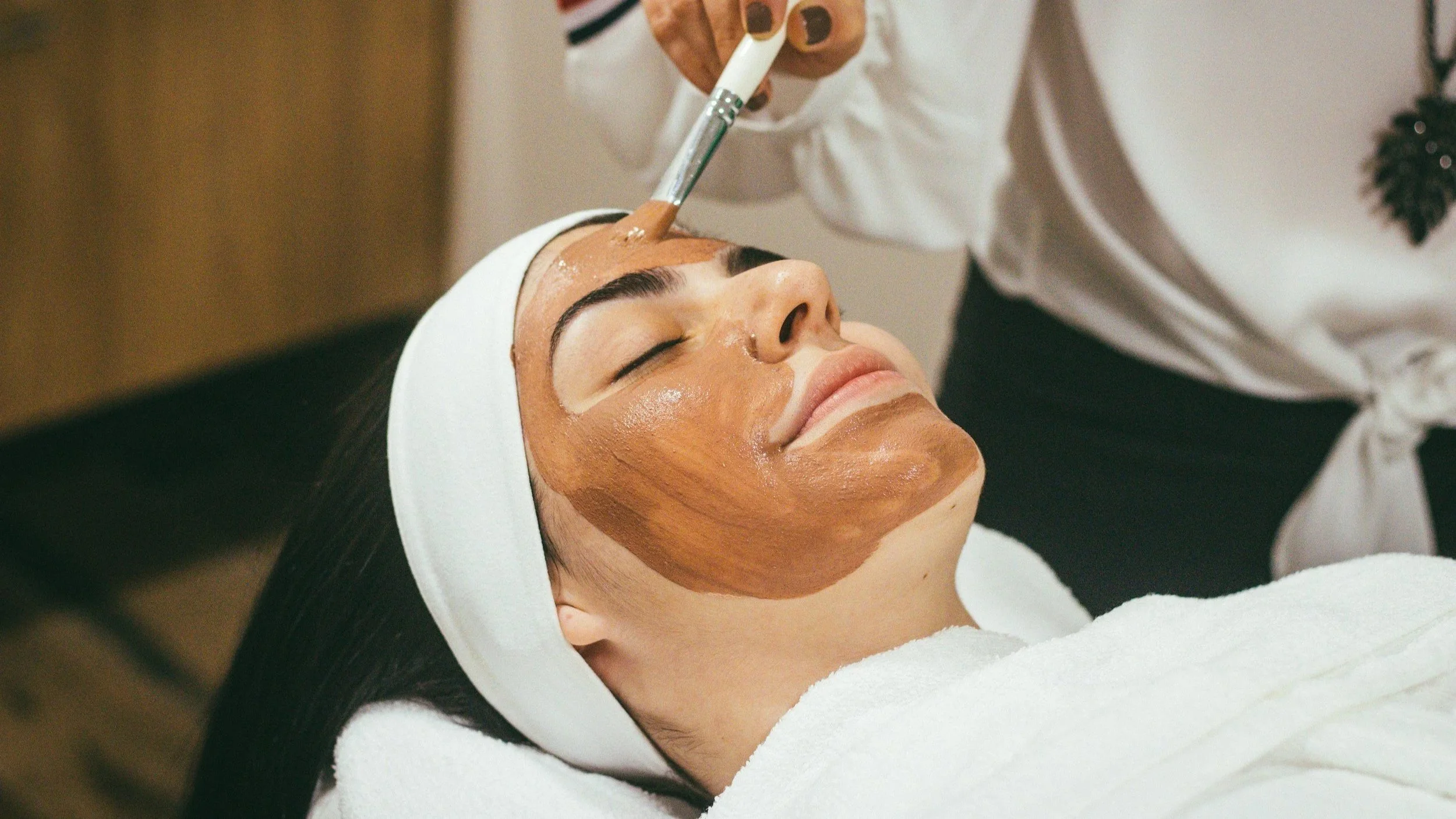 A woman lying down with her eyes closed, receiving a facial treatment with a brush applying a brown facial mask, wearing a white headband.