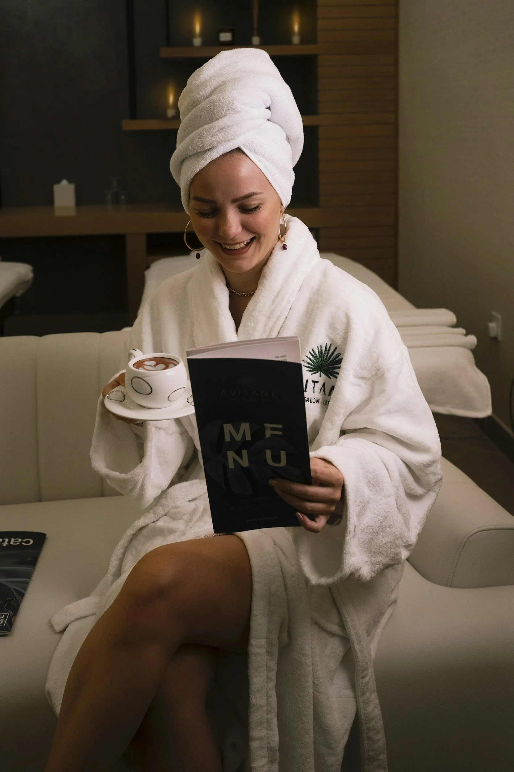 A woman wrapped in a white bathrobe and towel on her head, sitting on a couch, smiling while reading a menu. She has earrings and a necklace, and holds a cup of coffee.