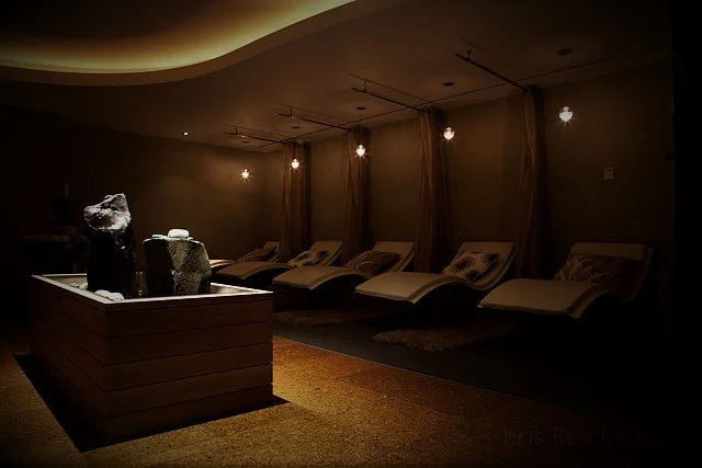A dimly lit room with six reclined lounge chairs, each with a pillow, and a wooden table with rock-like sculptures in the foreground.