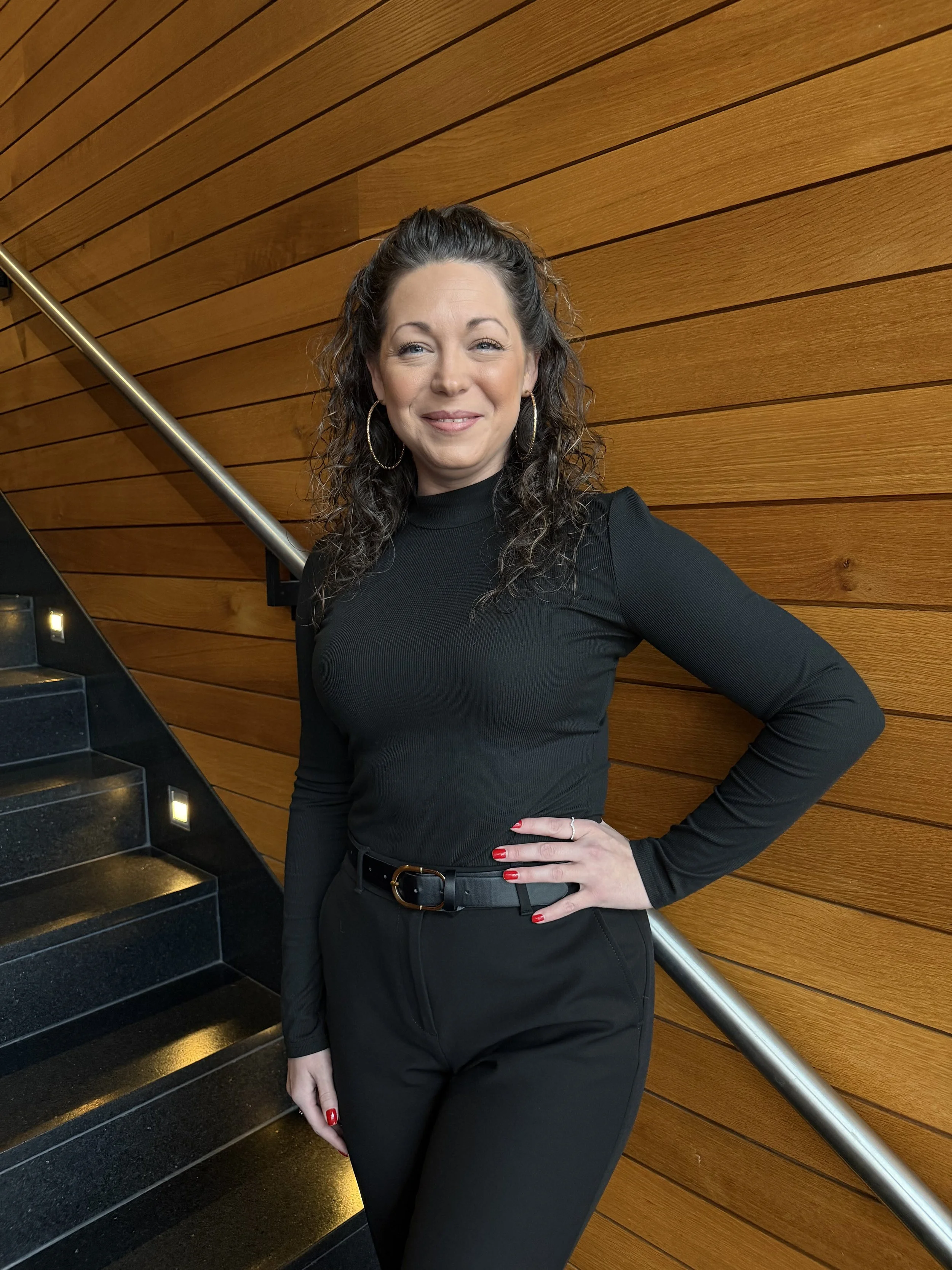 A woman with curly brown hair, wearing a black turtleneck and matching black pants, standing on a staircase with wooden paneling background.