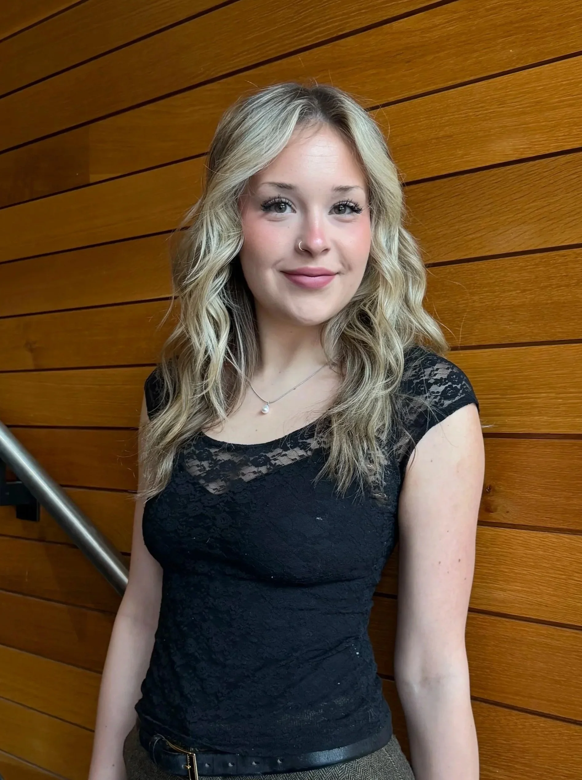 Portrait of a young woman with blonde, wavy hair, wearing a black lace top, a pearl necklace, and a small nose ring, standing in front of a wooden wall.