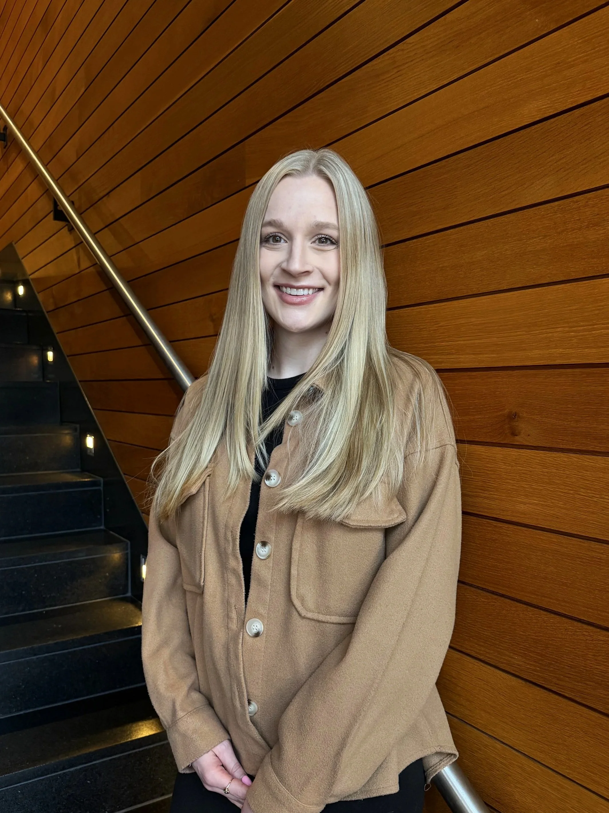A smiling woman with long blonde hair stands in front of a wooden wall and a staircase with black steps and built-in lighting.