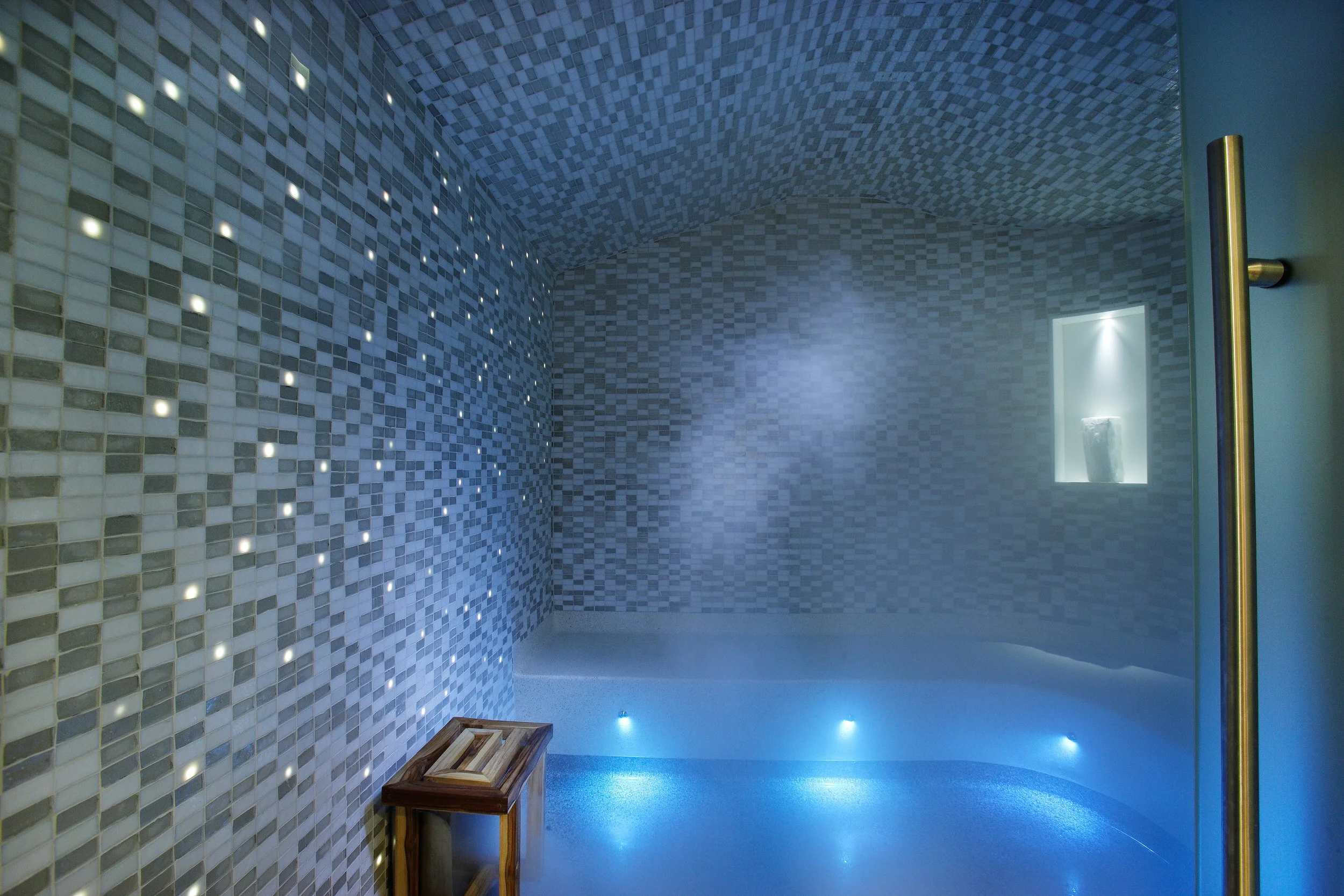 An indoor shower area with mosaic tile walls and ceiling, equipped with small embedded lights and a wooden bench.