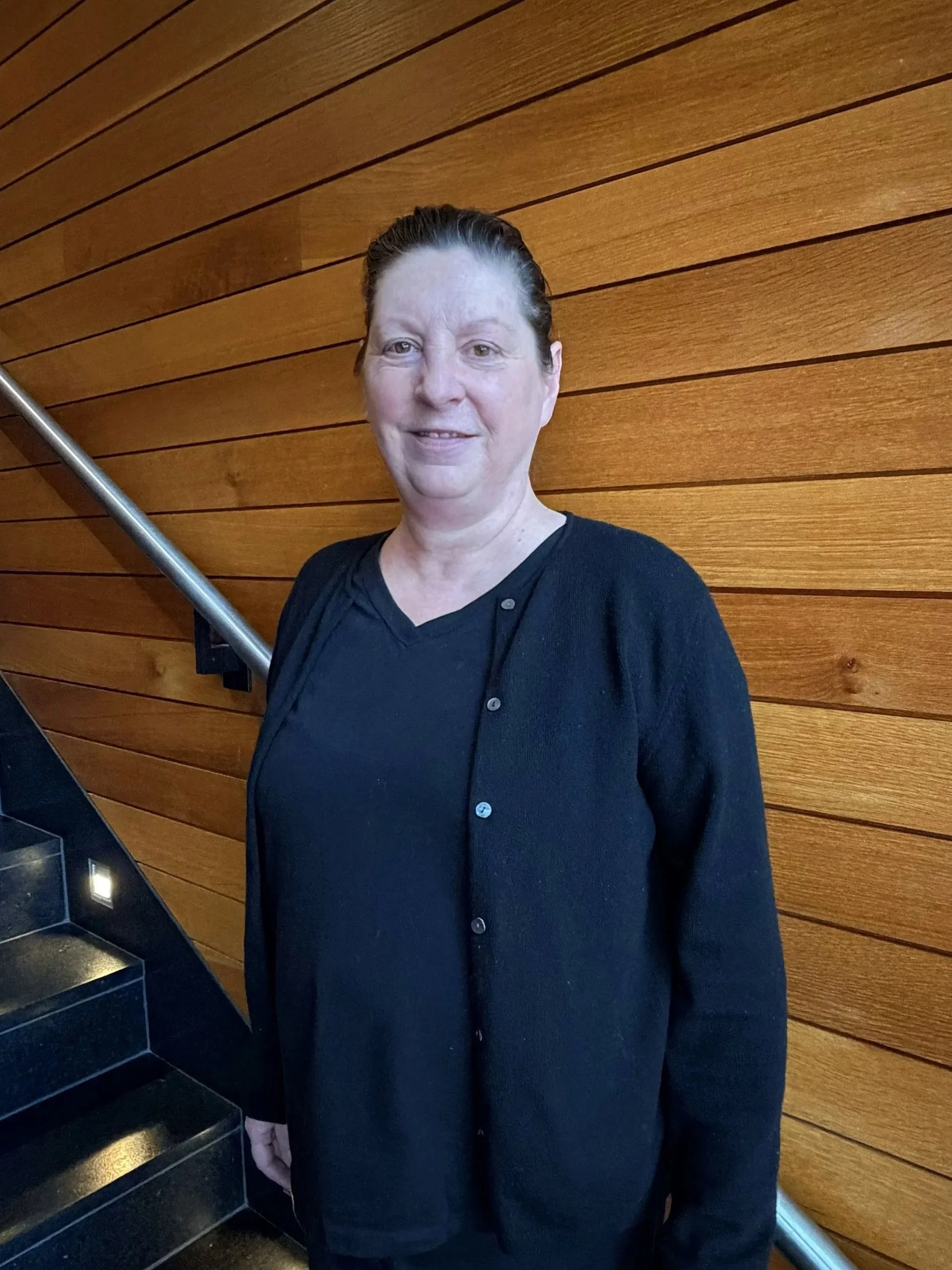 A woman standing indoors against a wooden wall, wearing a black shirt and cardigan, with dark hair pulled back, smiling.