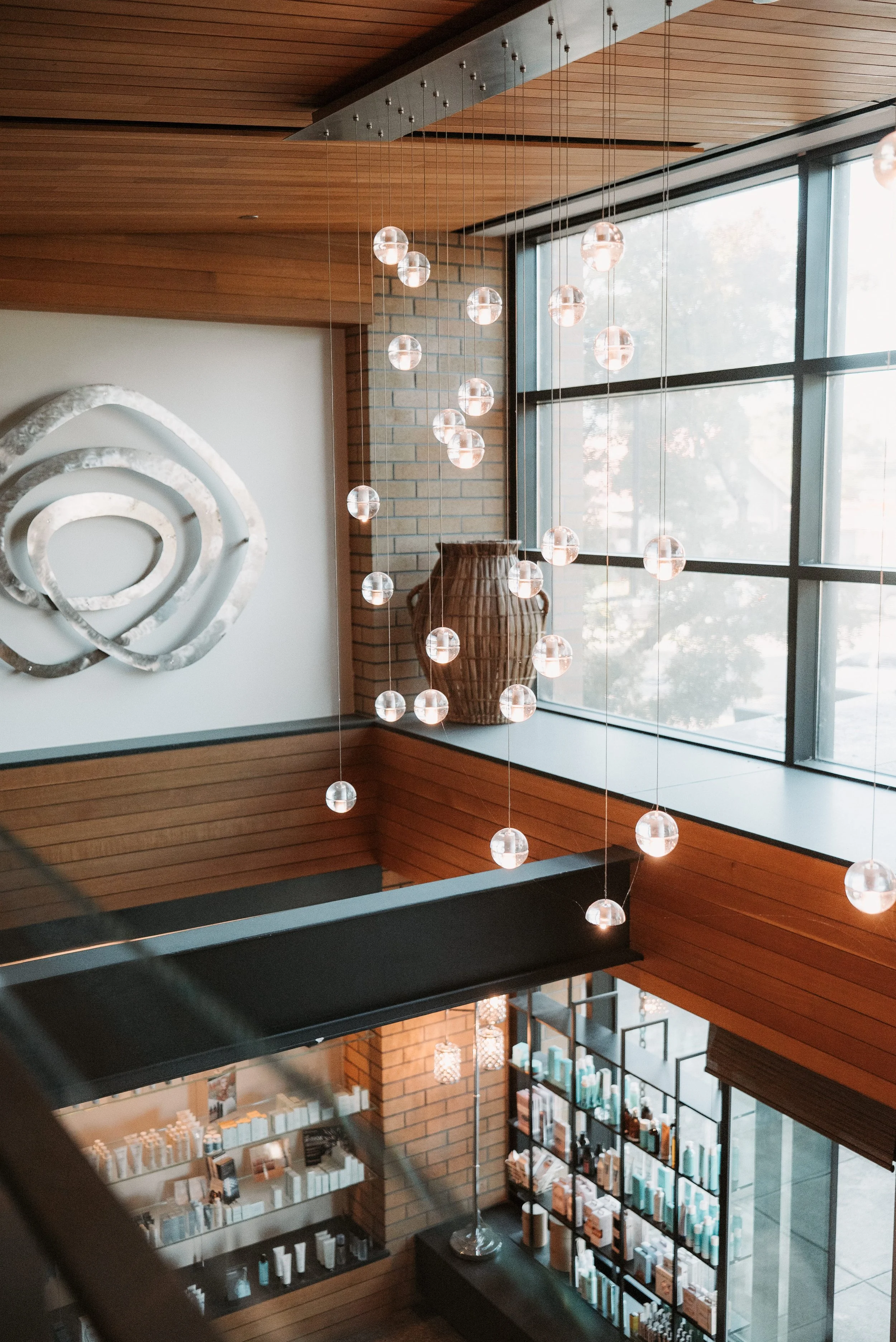 Interior of a modern building featuring hanging spherical pendant lights, a large window with a view outside, a decorative wall art piece with spiral design, a large woven vase, and shelves filled with books and decorative items.