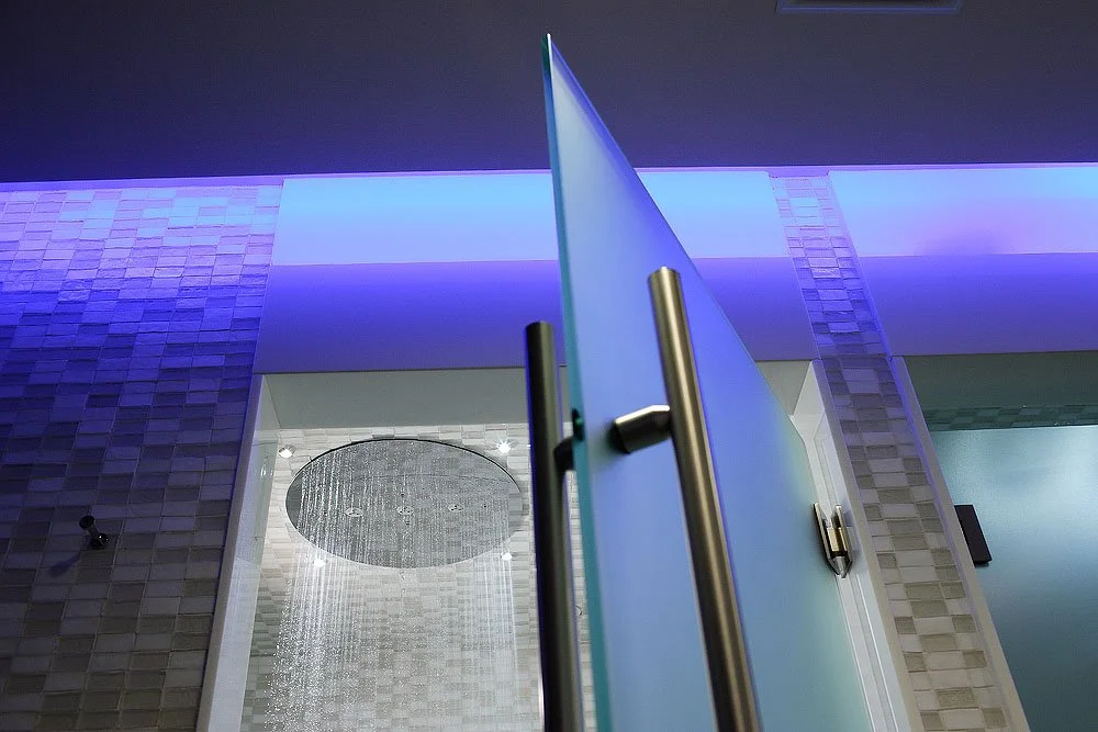 Close-up of a glass door with a metal handle in an interior space illuminated by blue and purple lighting, revealing a shower area with ceiling-mounted rainfall showerhead in the background.