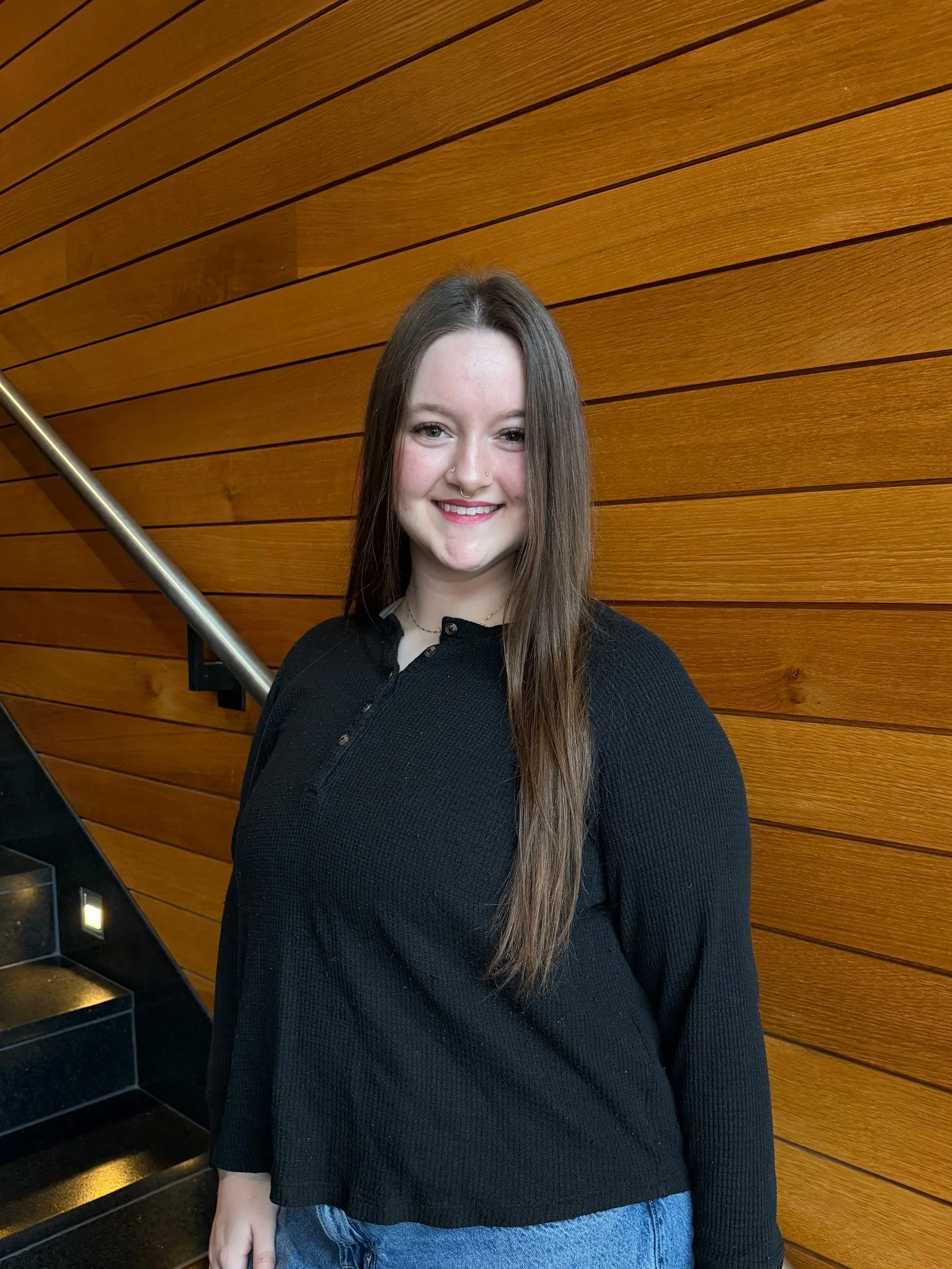 Young woman with long brown hair, smiling, wearing a black sweater and blue jeans, standing in front of a wooden wall with horizontal panels.