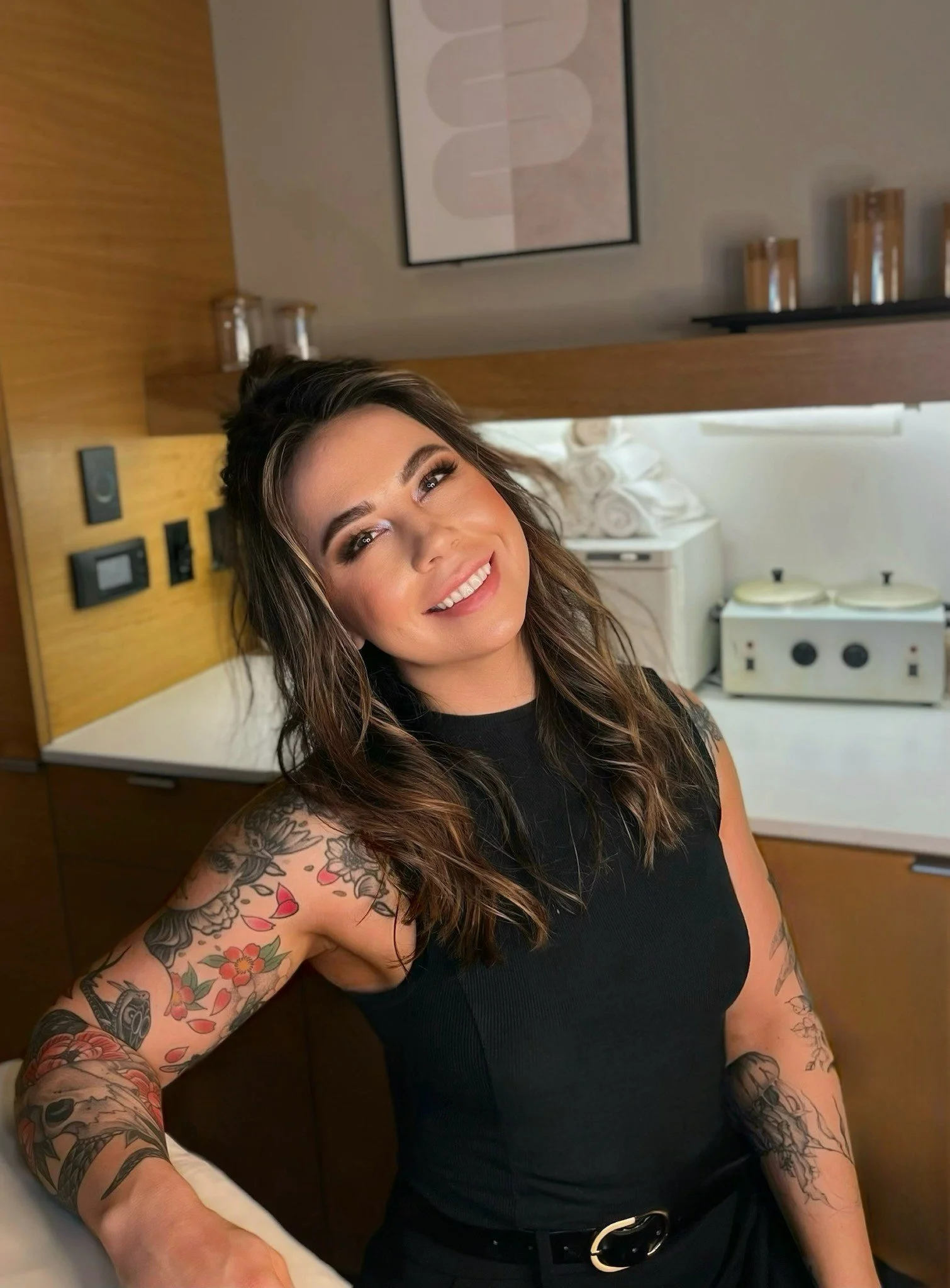 A woman with long, wavy brown hair and tattoos on her arm, smiling, wearing a black sleeveless top, sitting in a room with wooden cabinets and shelves, framed artwork on the wall, and some kitchen items in the background.