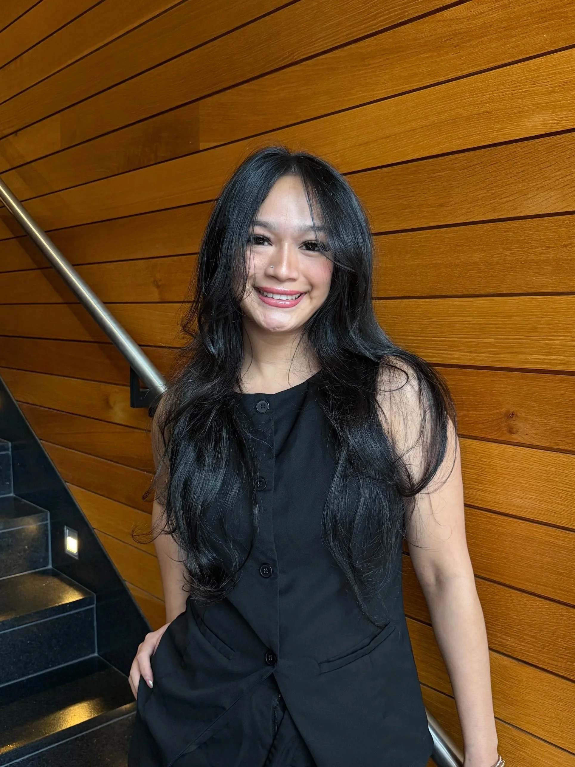 A woman with long black hair wearing a black sleeveless top and pants, smiling against a wooden wall background.
