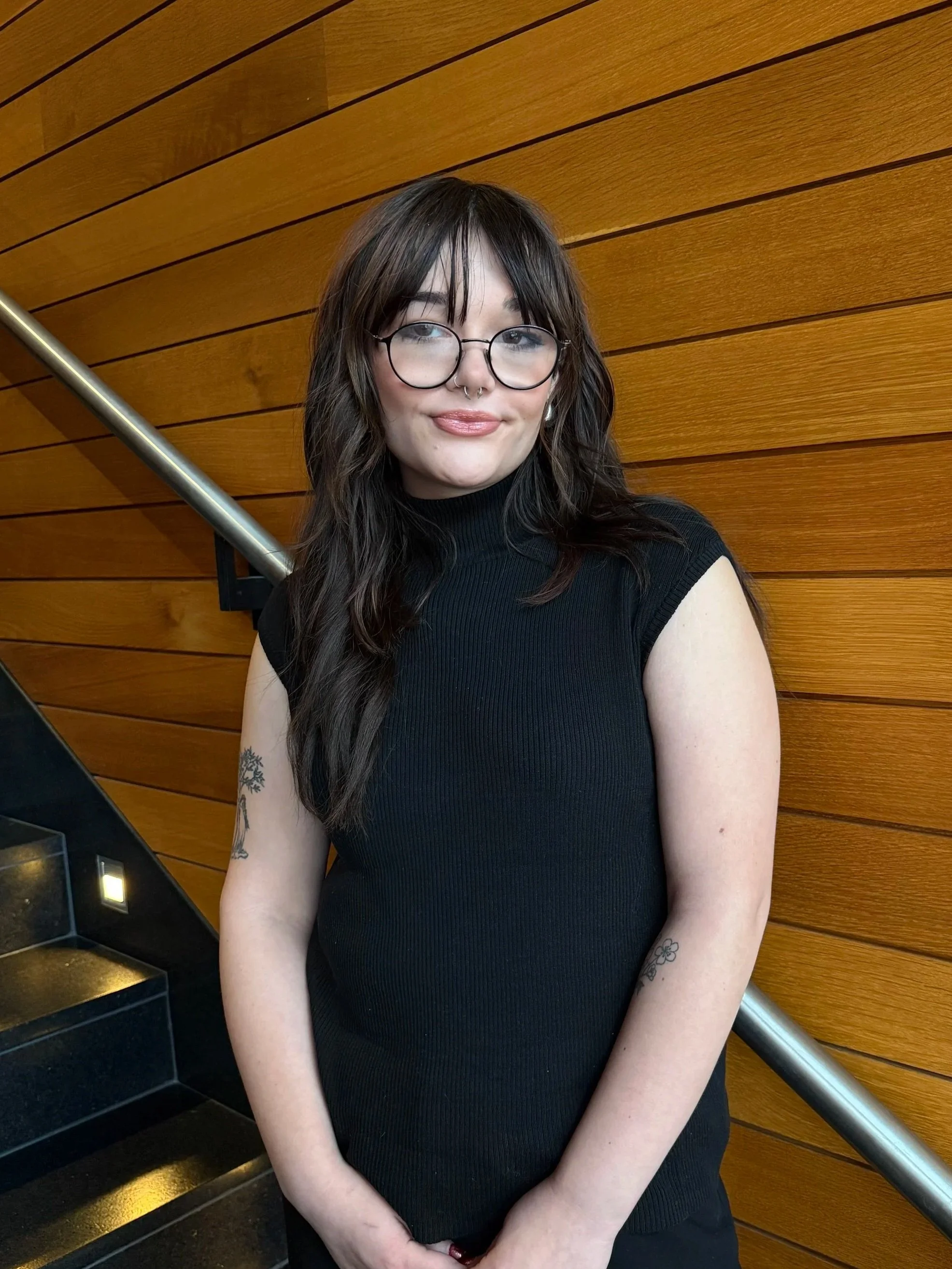 A young woman with long dark hair, wearing glasses and a black sleeveless top, is standing in front of wooden wall paneling with a metal handrail beside her. She has a slight smile and some tattoos on her arms.