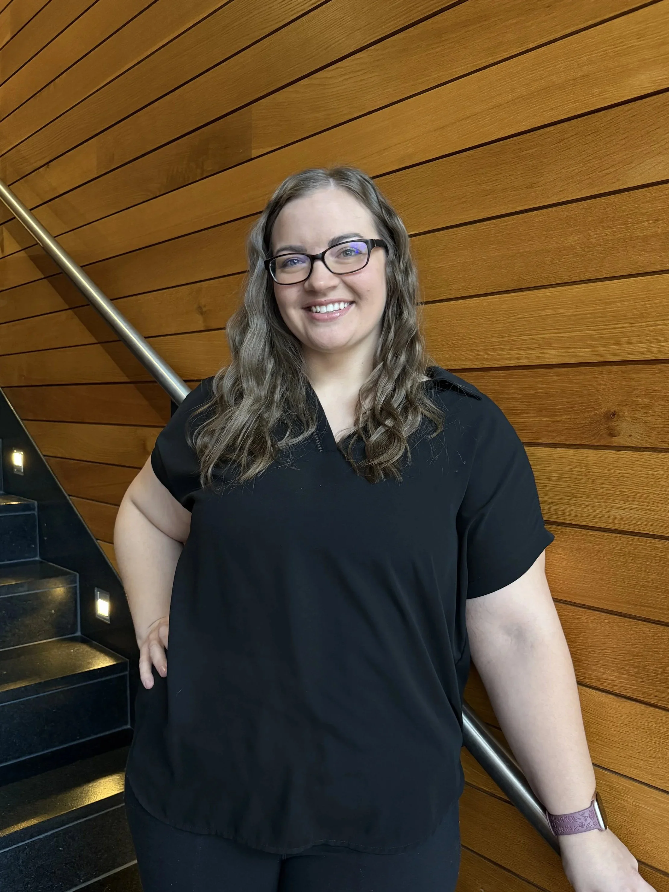 A smiling woman with glasses and long wavy hair wearing a black top, standing on a staircase with a wooden wall in the background.