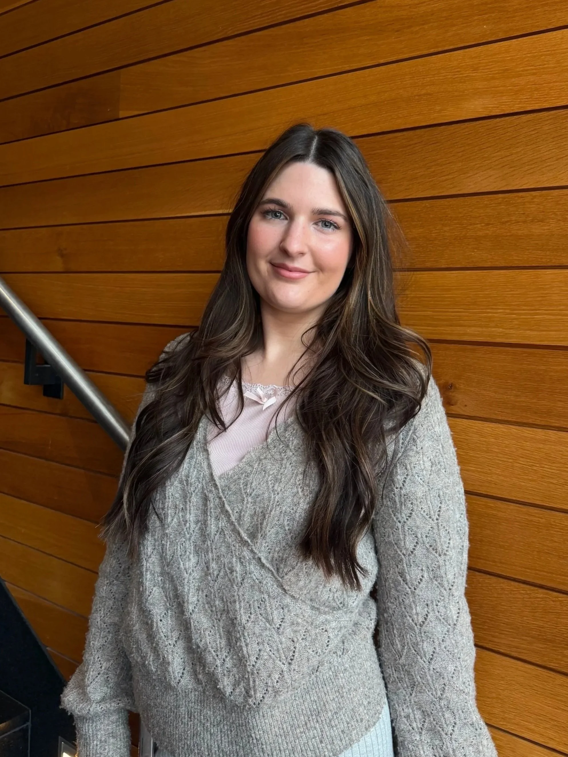 A woman with long brown hair and blue eyes standing in front of a wooden wall, wearing a gray cable knit sweater.