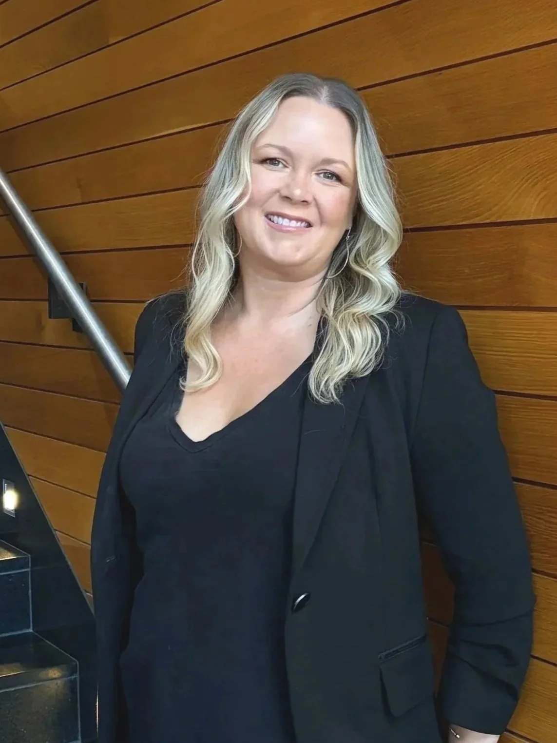 A woman with blonde, wavy hair wearing a black blazer and black top, standing against a wooden-paneled wall with a metal handrail on the left.