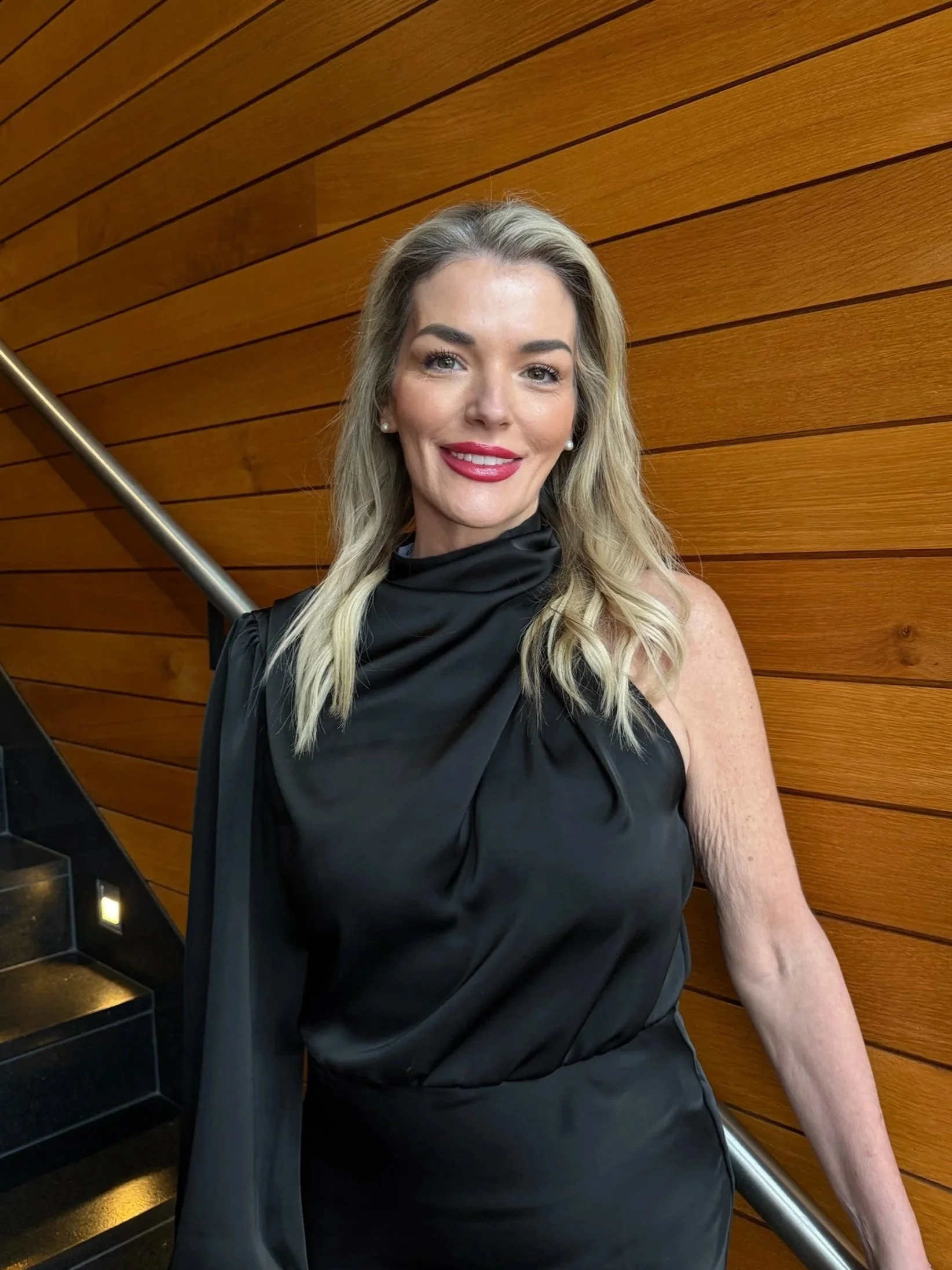 A smiling woman with long blonde hair, wearing earrings, a black dress with a draped neckline, and red lipstick. She is standing beside a staircase with a wooden wall in the background.