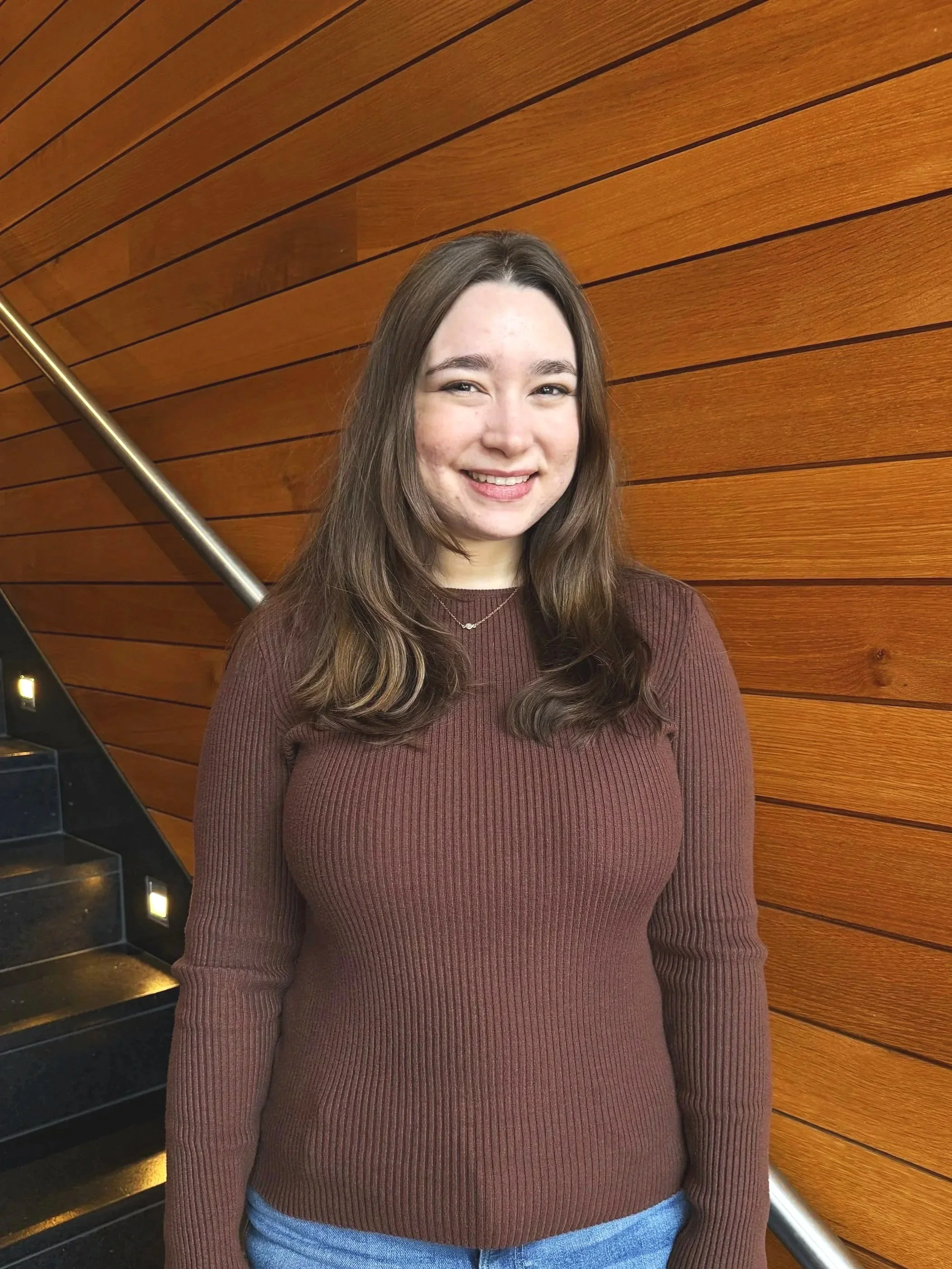 A young woman with long brown hair, wearing a maroon ribbed sweater and blue jeans, standing in front of a wooden wall with horizontal planks and a metal handrail.
