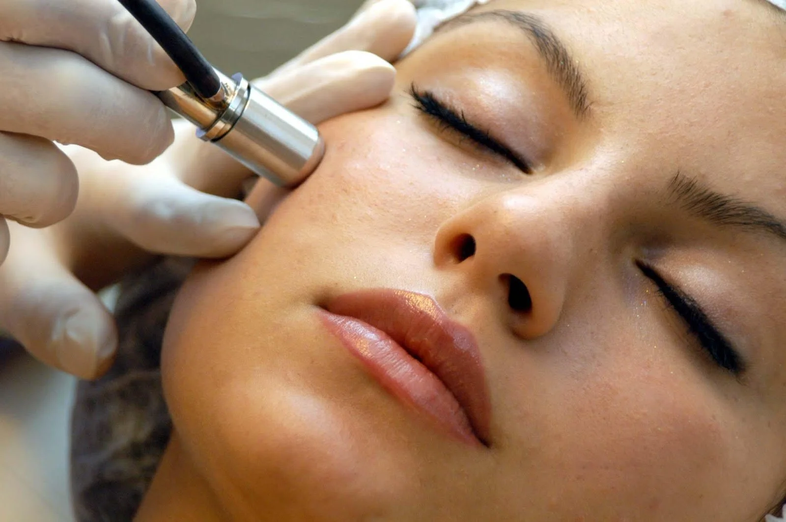 A woman receiving a cosmetic or skincare treatment on her face by a professional wearing gloves, using a small device near her cheek.