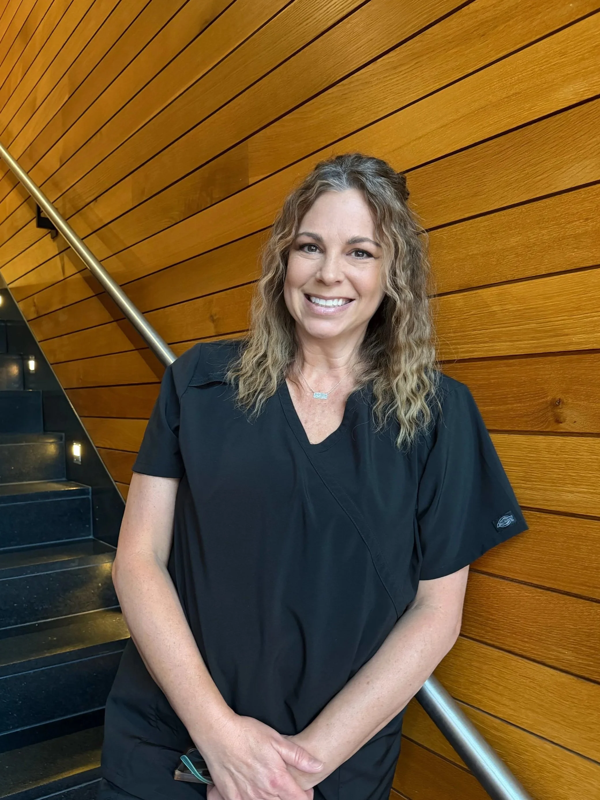A woman with curly hair smiling, wearing a navy blue shirt, standing on a staircase with black steps and small lights, against a wooden wall with horizontal panels.