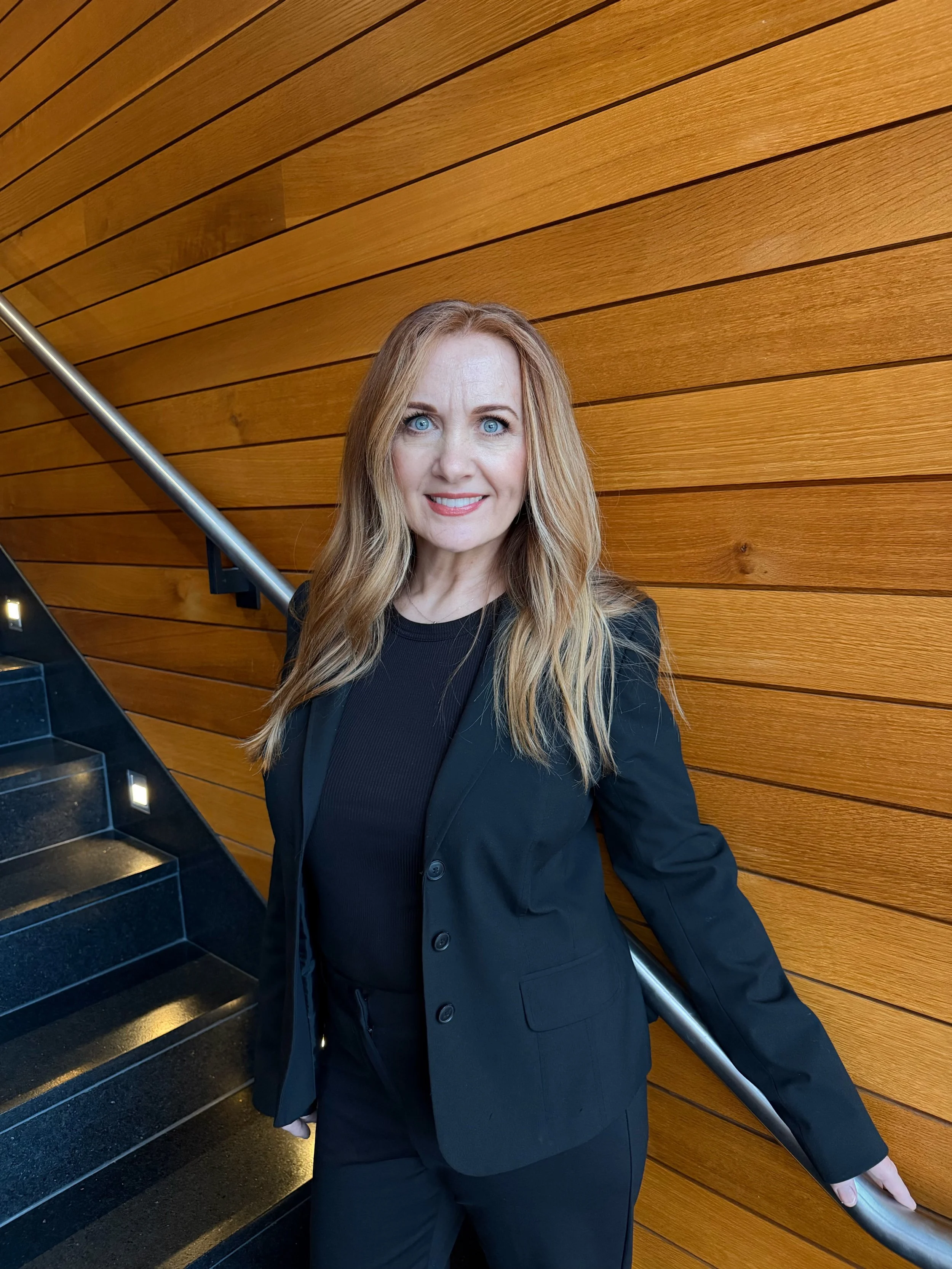 A woman with long blonde hair and blue eyes standing near black stairs and a wooden wall, smiling at the camera.