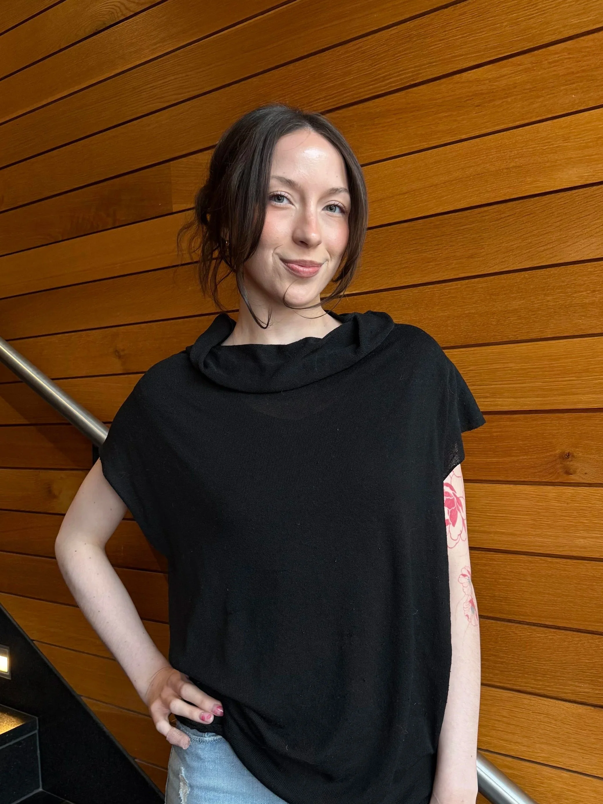 A young woman with shoulder-length brunette hair, blue eyes, and fair skin is standing indoors against a wooden wall with horizontal paneling. She is wearing a black top and light blue jeans, with her right hand on her hip, smiling softly at the camera.
