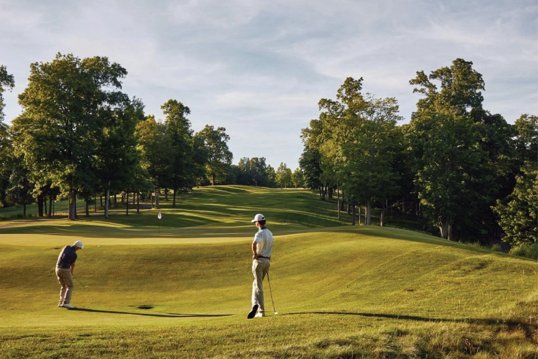 Two golfers on a golf course near the hole, surrounded by green grass and trees with a bright sky overhead.