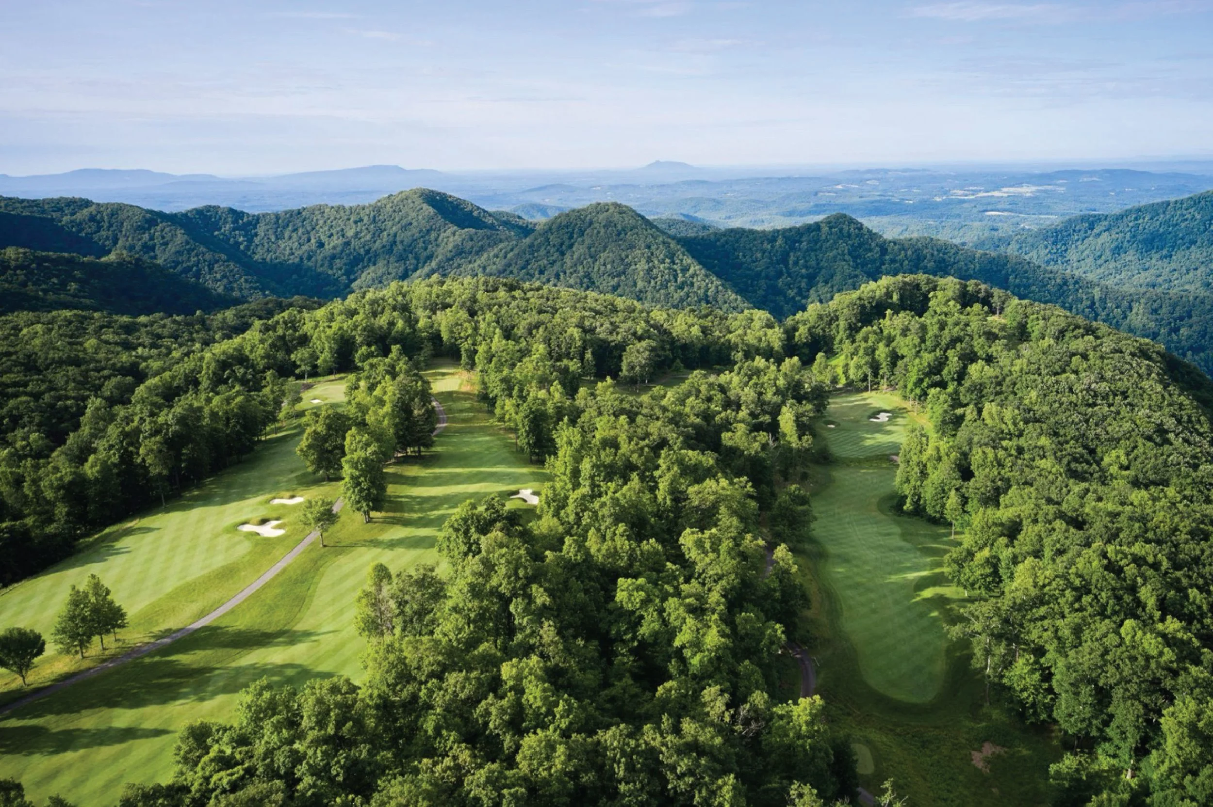 Aerial view of a lush green golf course with sand traps, surrounded by dense forest and mountains in the background.