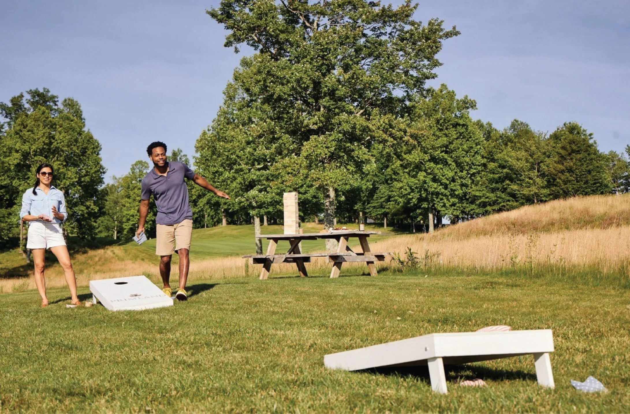 Two people playing cornhole in a park with grass, trees, a picnic table, and blue sky.