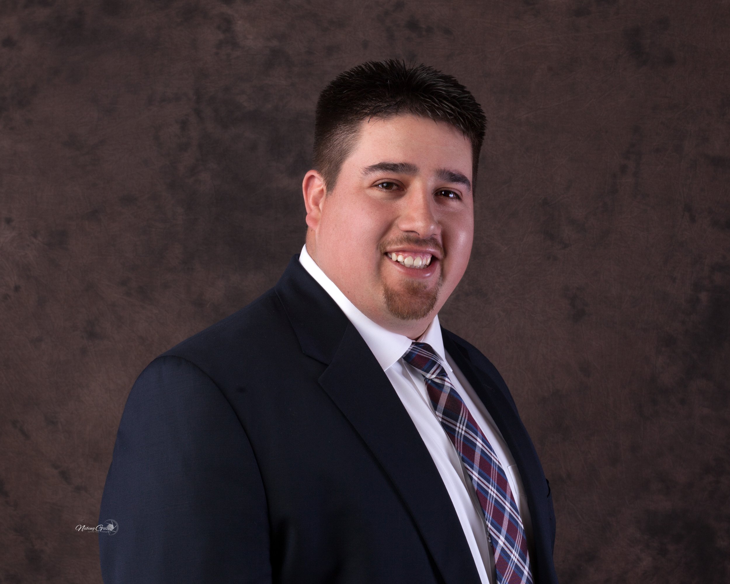 A man in a dark suit, white dress shirt, and plaid tie smiling at the camera against a brown textured background.