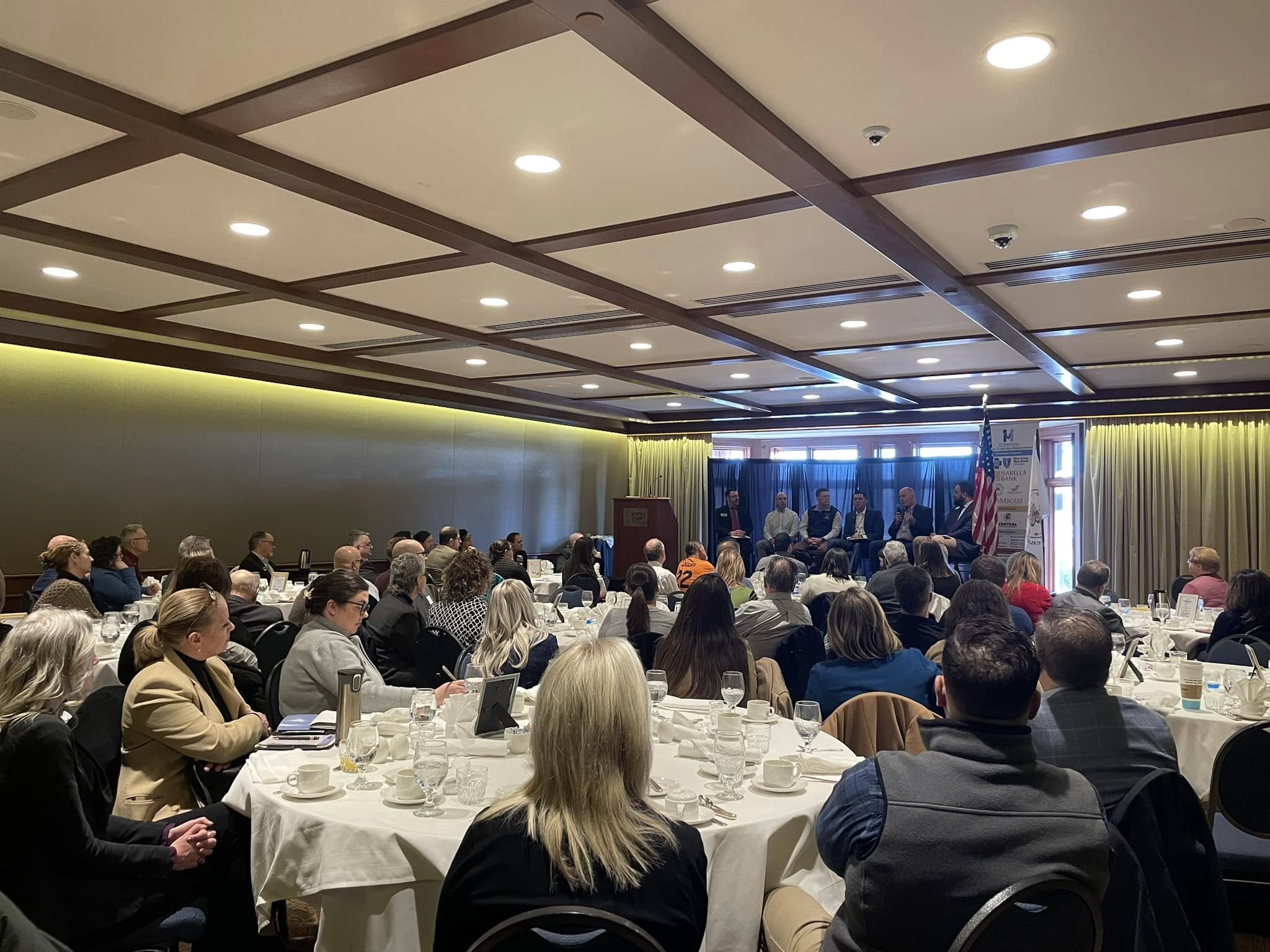 A large conference room filled with attendees sitting at round tables, listening to a panel of speakers seated on a stage at the front, with a U.S. flag and a banner nearby.