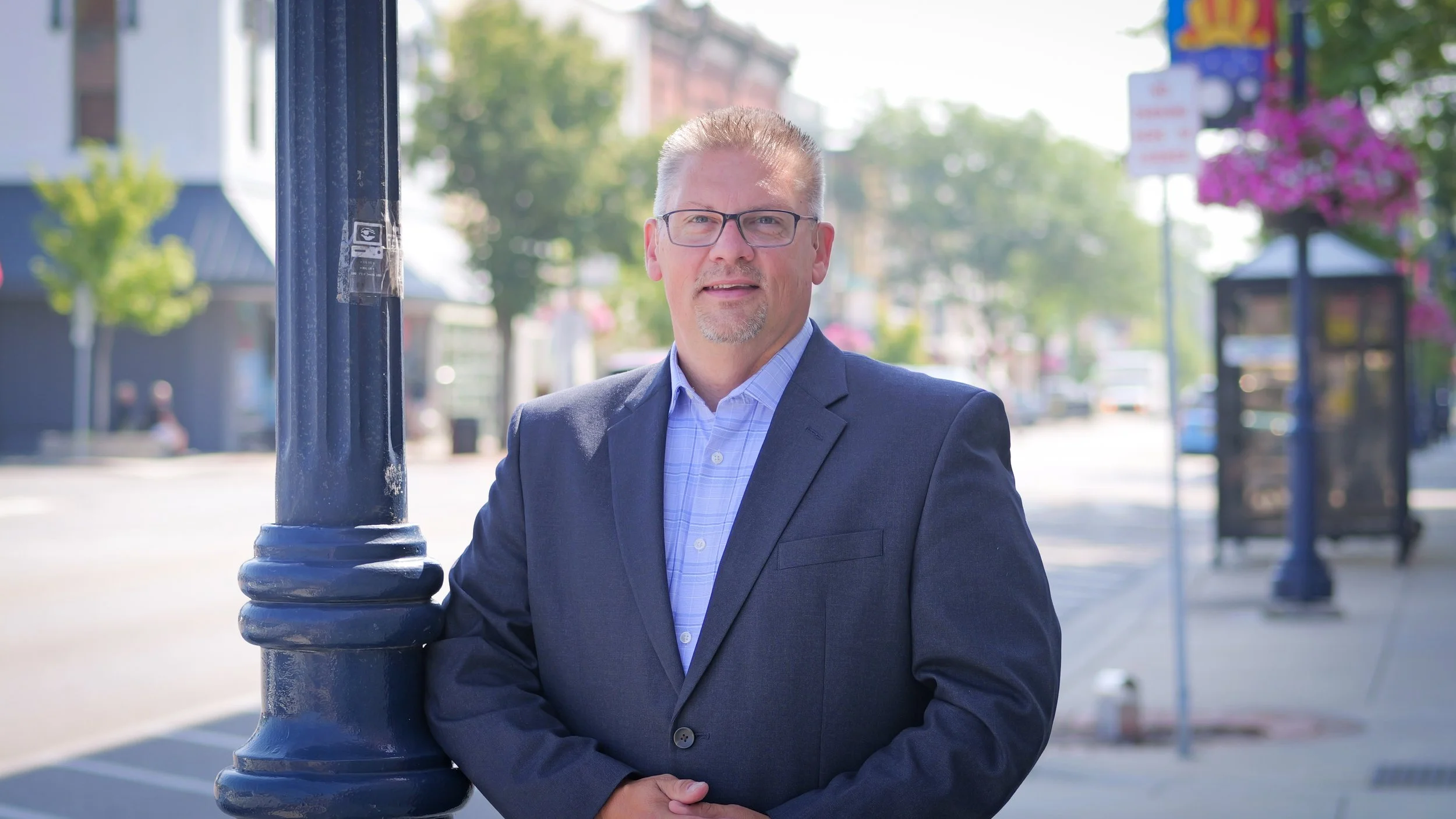 A man in a dark suit and glasses standing outdoors next to a street lamp on a city street.