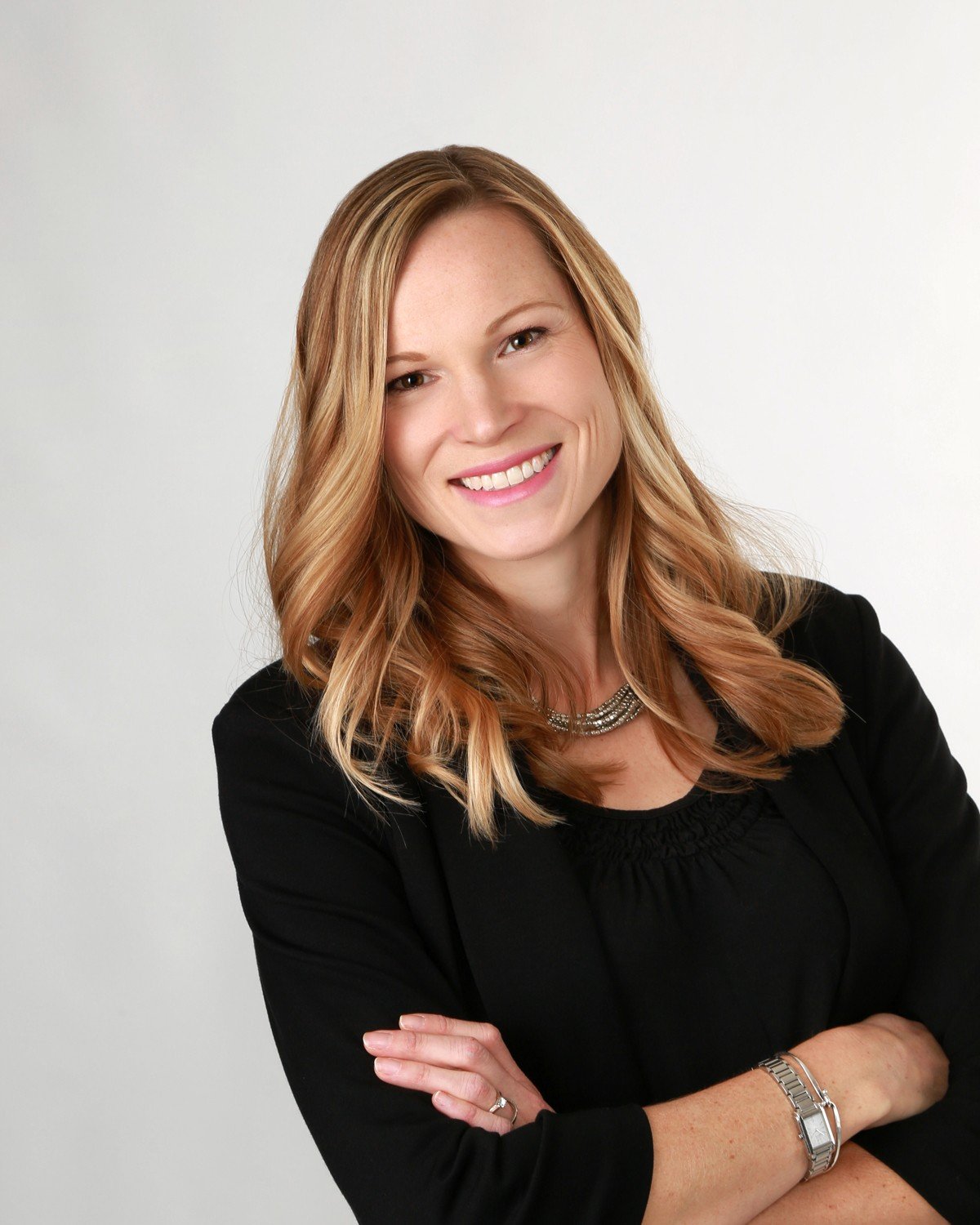 Head and shoulders portrait of a smiling woman with light brown hair, crossed arms, wearing a black blazer, silver necklace, and watch, against a plain light background.
