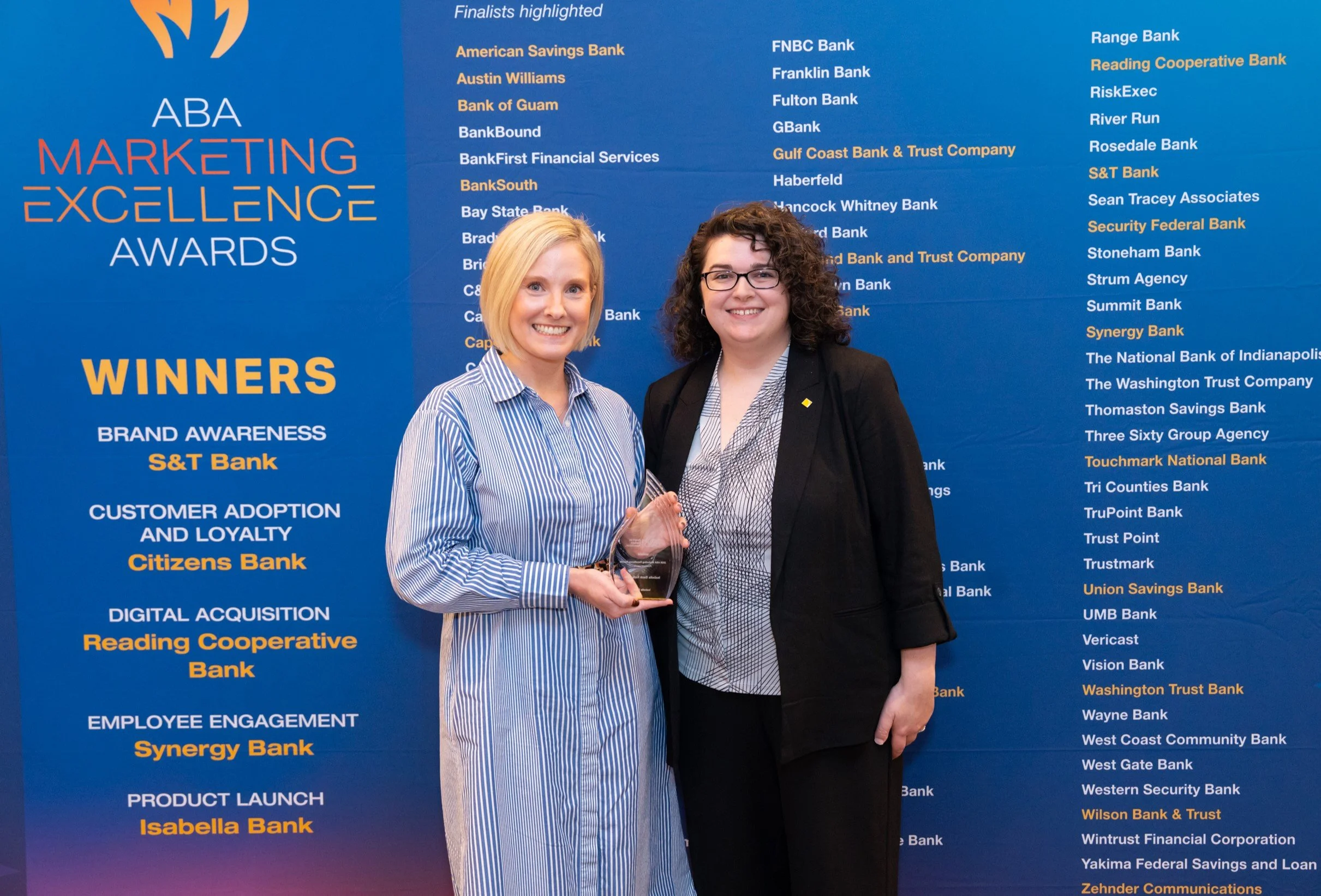 A woman with short blonde hair and a woman with short brown curly hair standing in front of a blue backdrop holding a glass award.