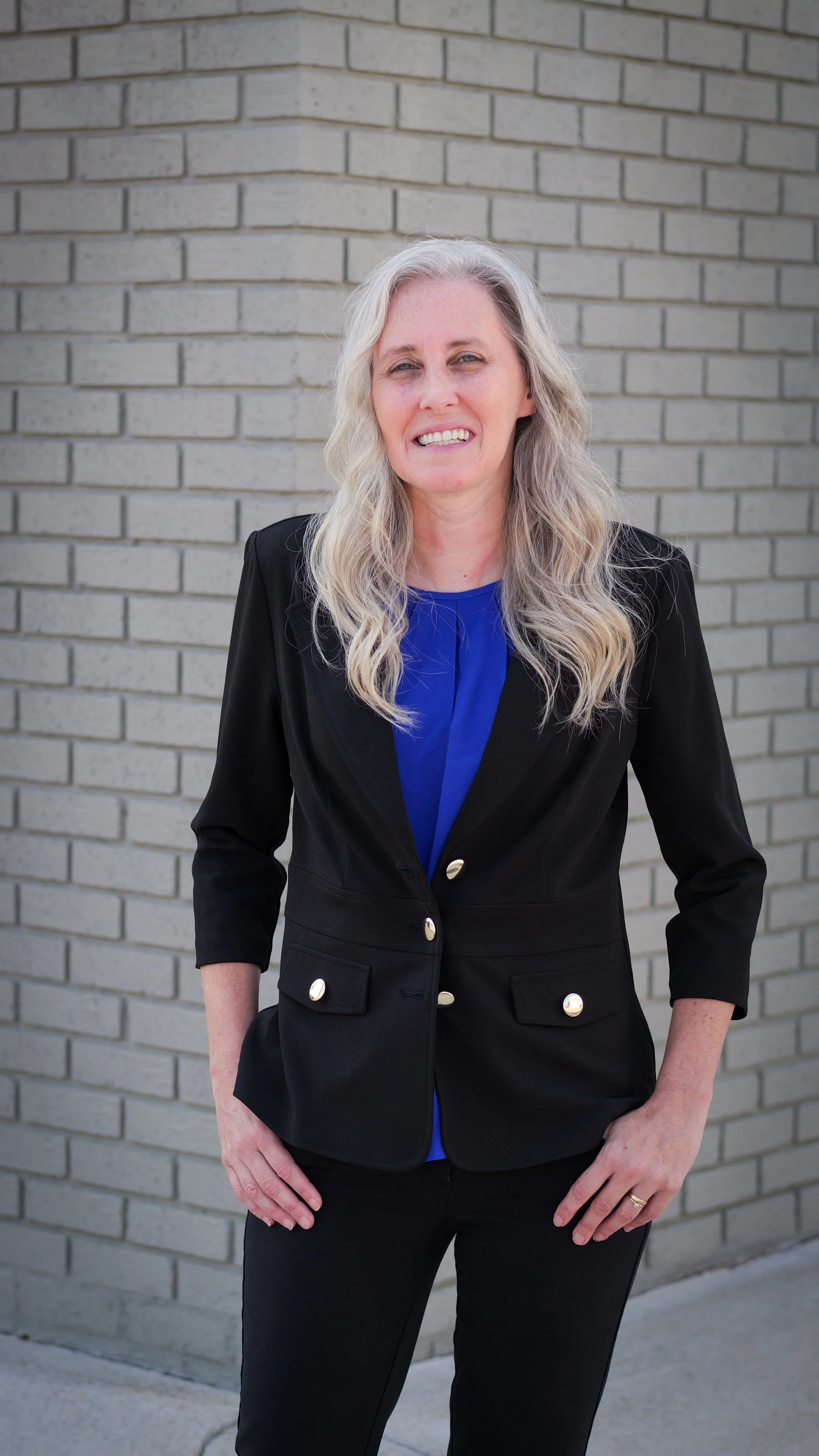 A woman with long, wavy gray hair stands outdoors in front of a gray brick wall. She is wearing a black blazer with gold buttons, a blue blouse, and black pants. She is smiling slightly and has one hand on her hip.