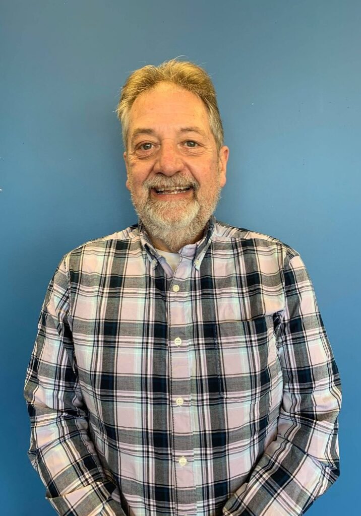 A middle-aged man with gray hair and a beard, smiling, wearing a plaid button-up shirt, standing against a blue wall.