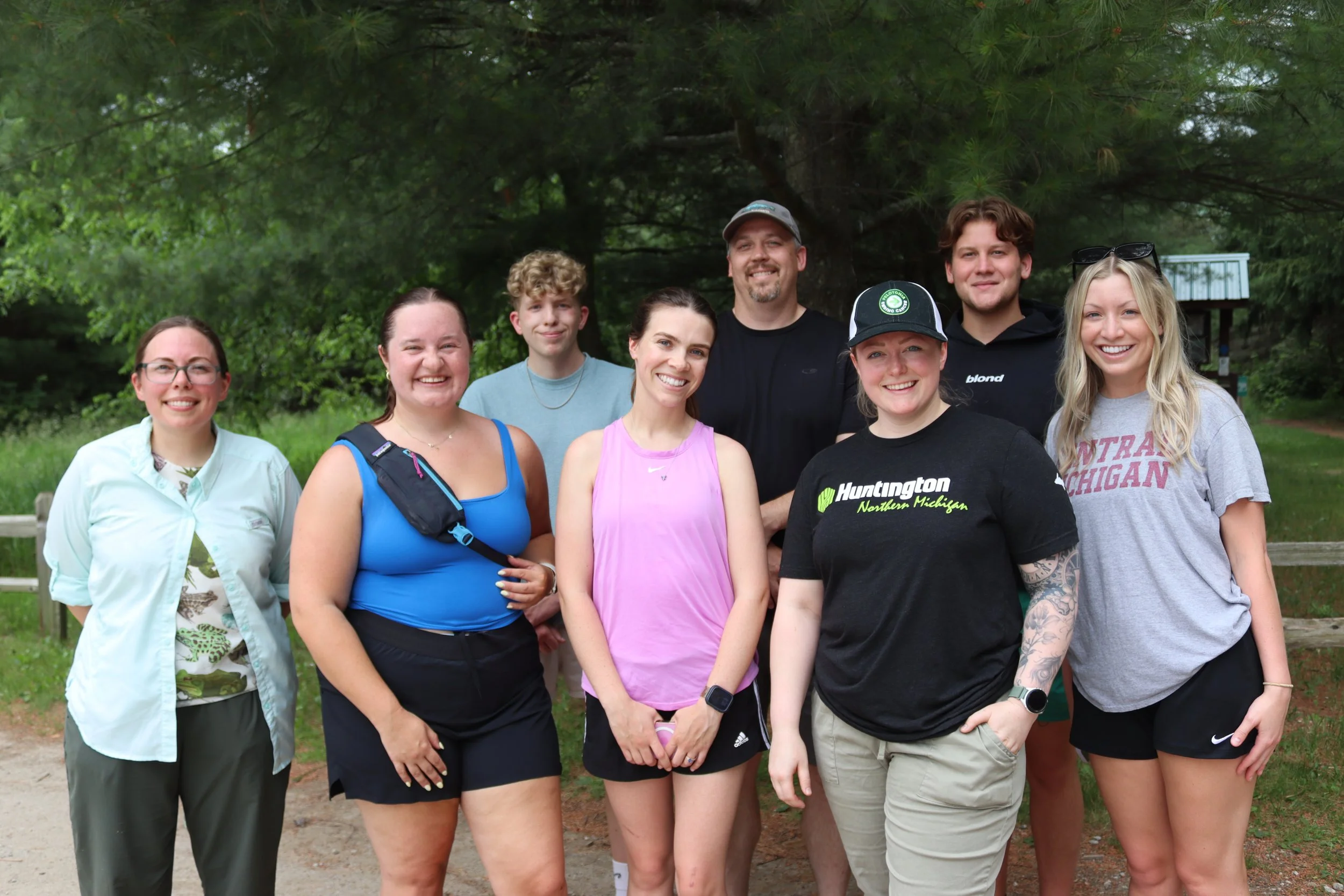 A group of nine young adults standing outdoors in front of green trees, smiling for a group photo.