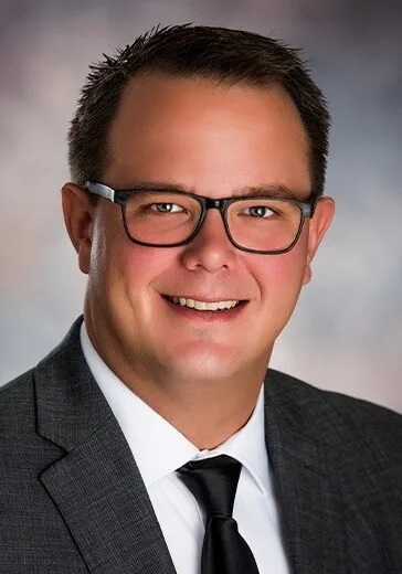 A man with short dark hair, glasses, and a broad smile, wearing a dark suit, white shirt, and black tie, posing in front of a neutral background.