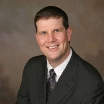 Headshot of a man in a black suit with a black tie against a brown background smiling.