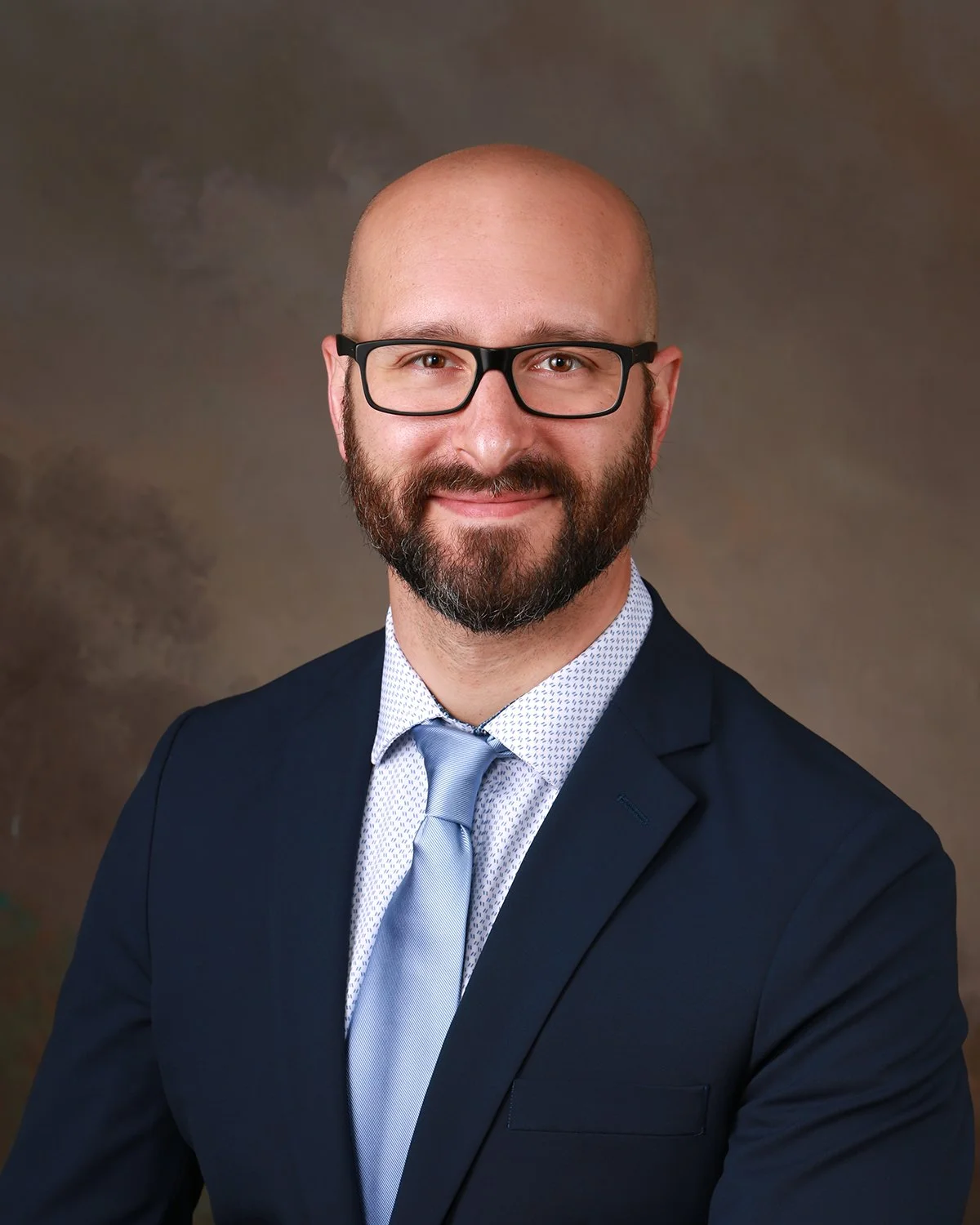 A professional headshot of a man with glasses, a beard, and a bald head, dressed in a navy suit, light blue shirt, and matching tie, smiling against a brown background.