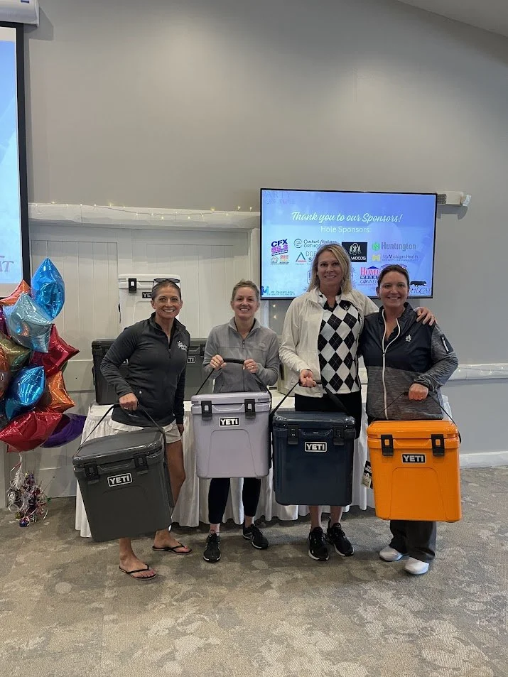 Four women standing indoors, smiling, each holding YETI cooler. Behind them is a large screen displaying sponsor logos and a thank you message. To the left, there are colorful balloons.
