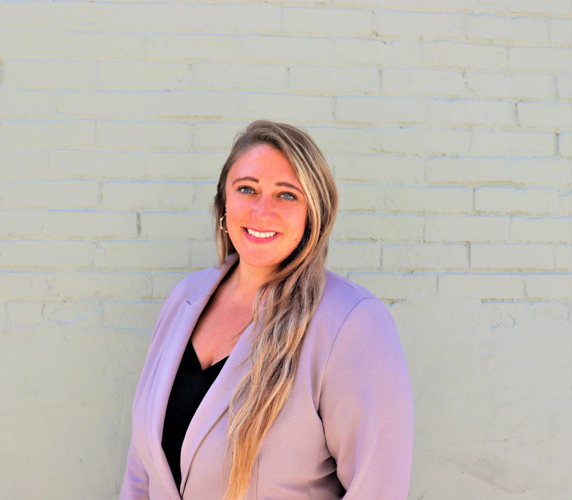A woman standing in front of a white brick wall smiling with a light purple blazer
