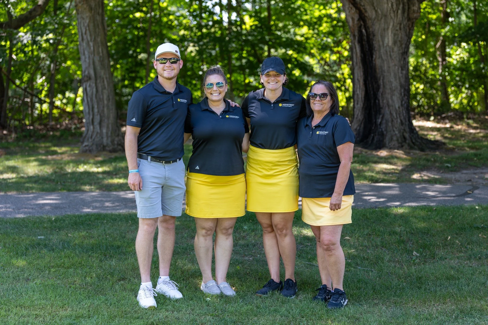 Four people standing together outdoors in front of trees, dressed in matching navy shirts with yellow skirts or shorts, smiling at the camera.