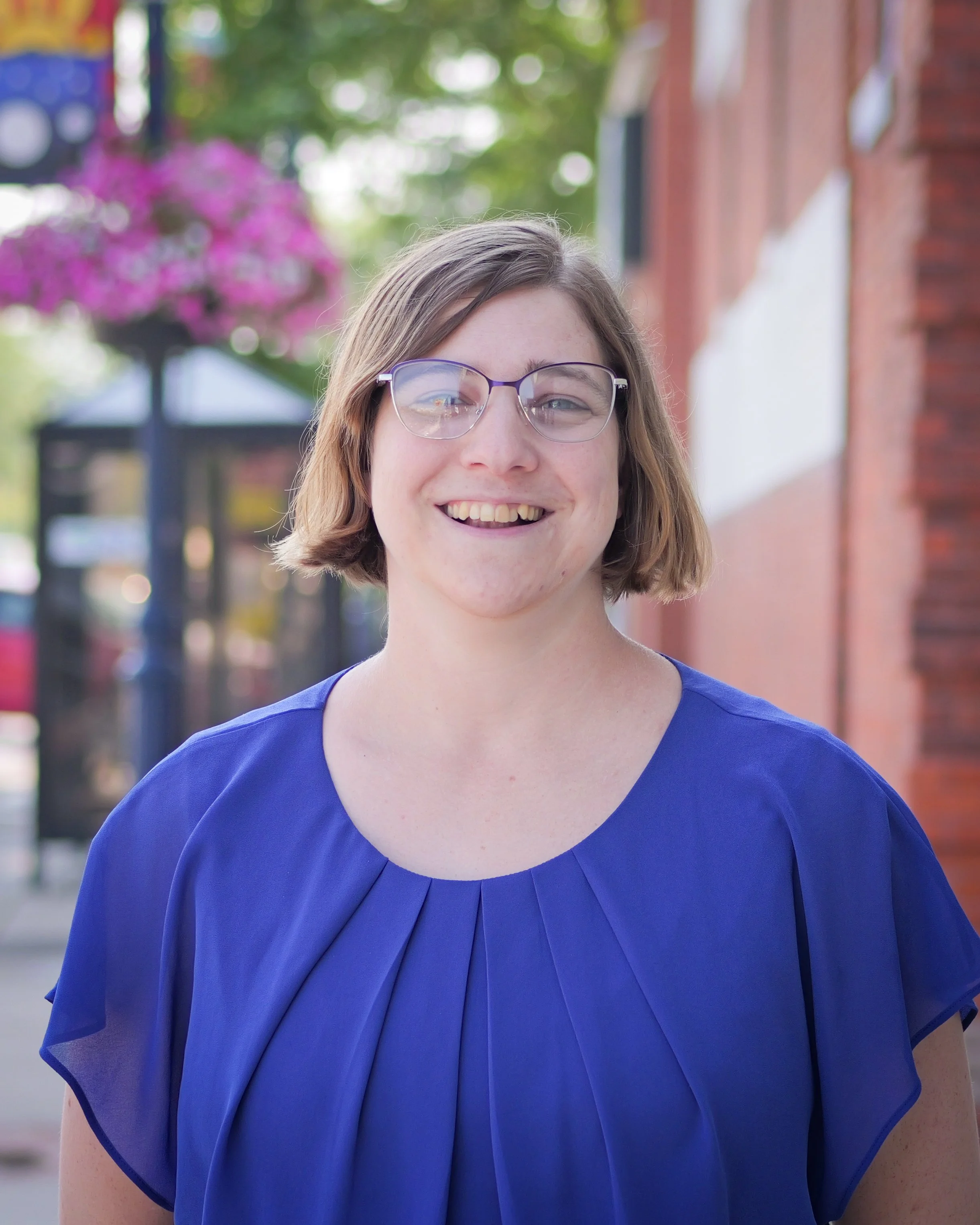A young woman with short light brown hair, glasses, and a bright smile, standing outdoors on a sidewalk with pink flowers and a brick building in the background, wearing a royal blue blouse.