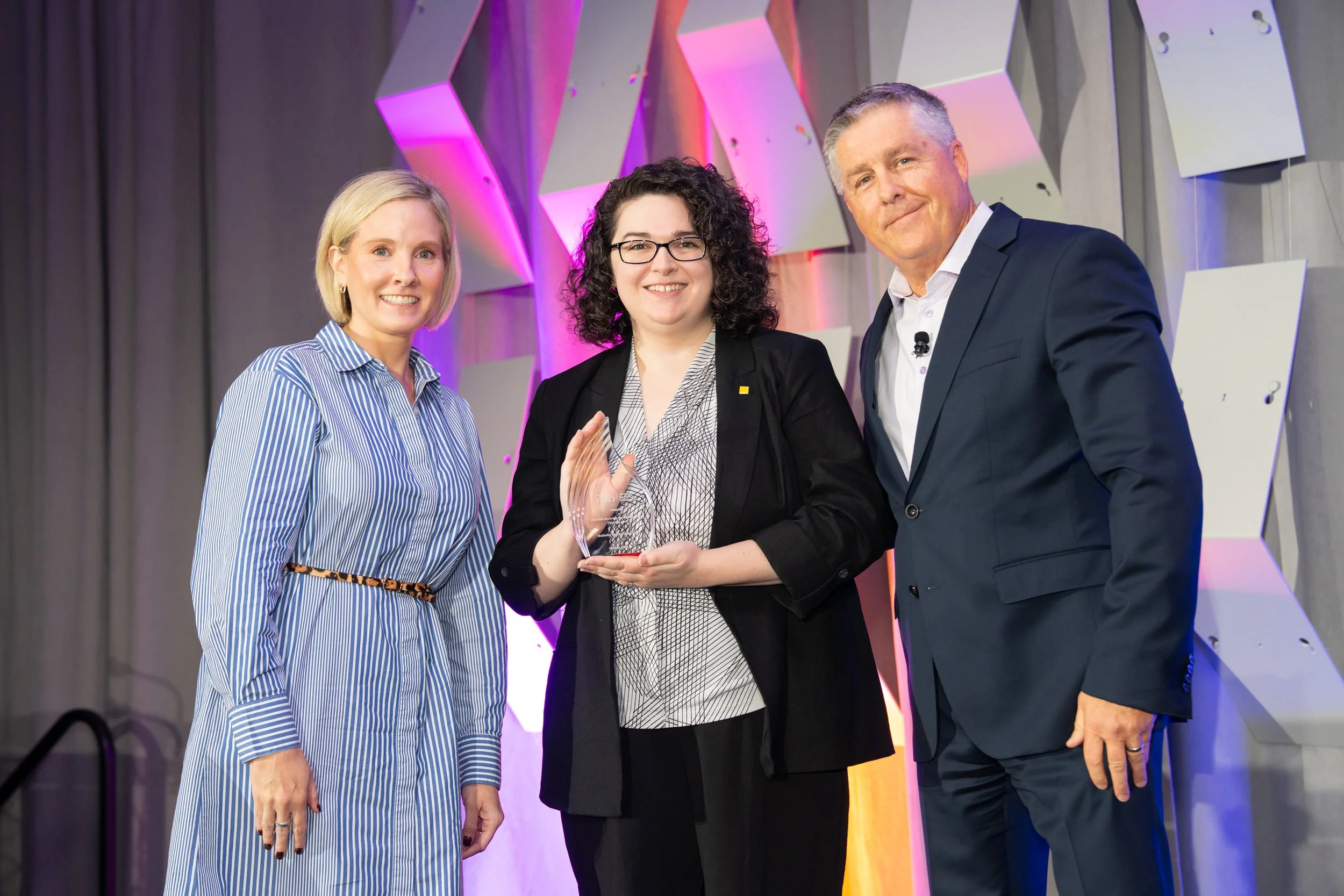 Woman with short blonde hair, woman with short curly hair and man with short gray hair standing together smiling while holding a clear, glass award