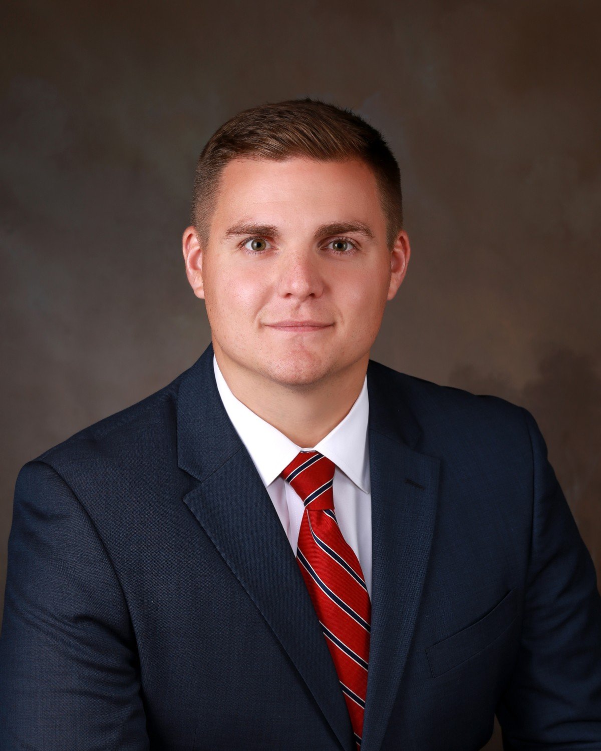 Professional portrait of a young man in a navy blue suit, white shirt, and red striped tie against a neutral background.
